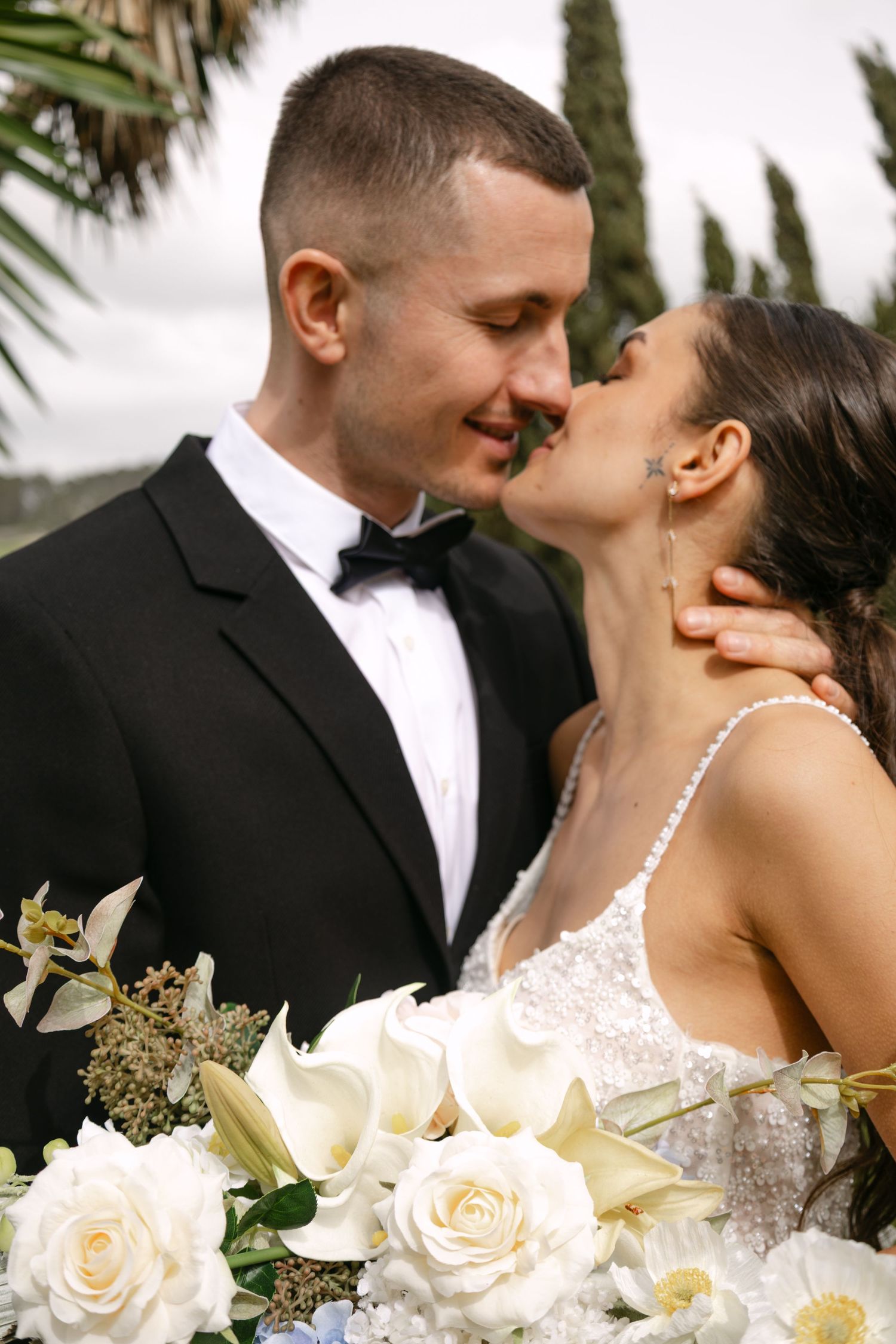 A bride and groom share a romantic kiss outdoors, with the bride holding a bouquet of white roses and lilies.
