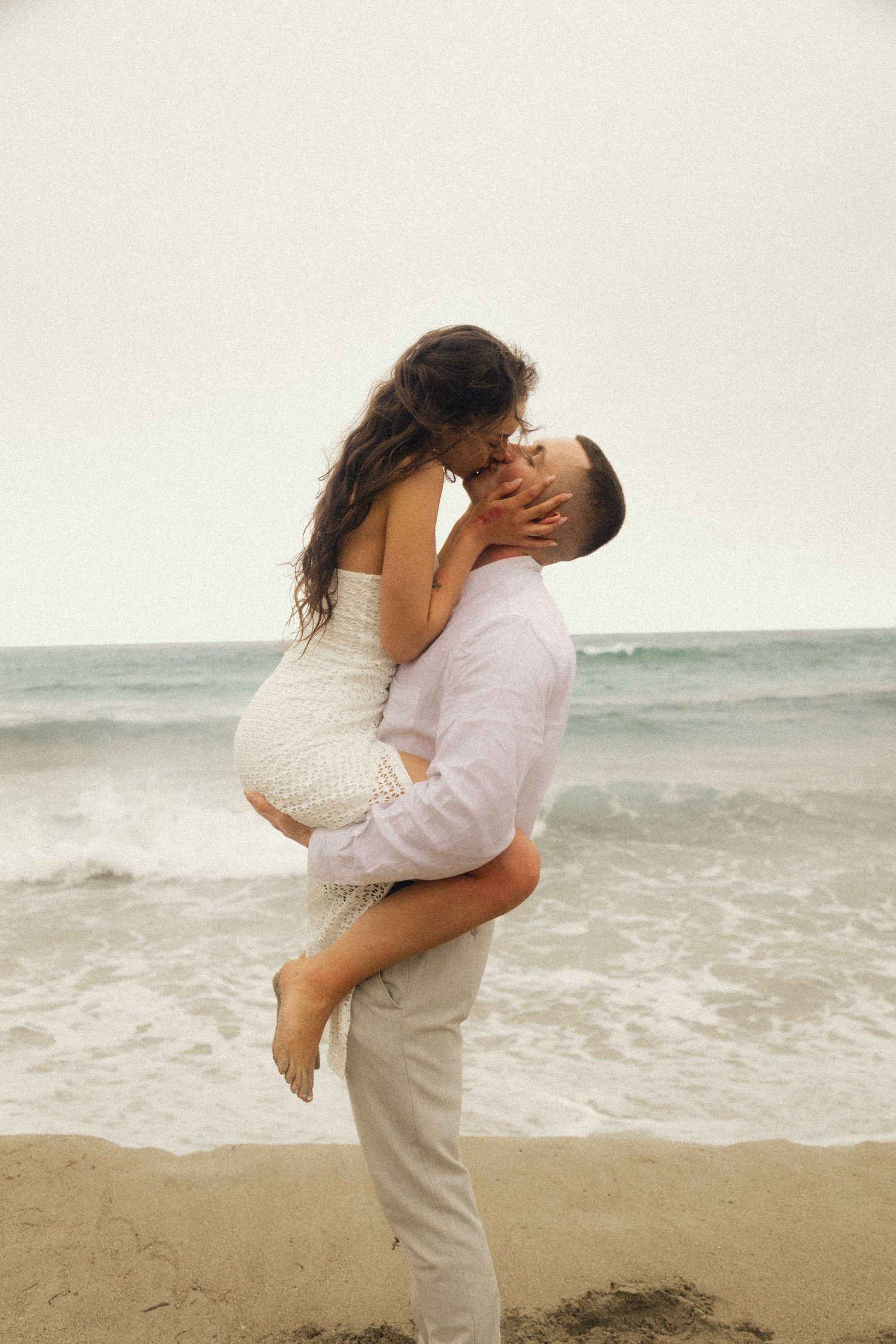 Romantic couple embracing on a sandy beach, man lifting woman in white dress with ocean waves behind them.