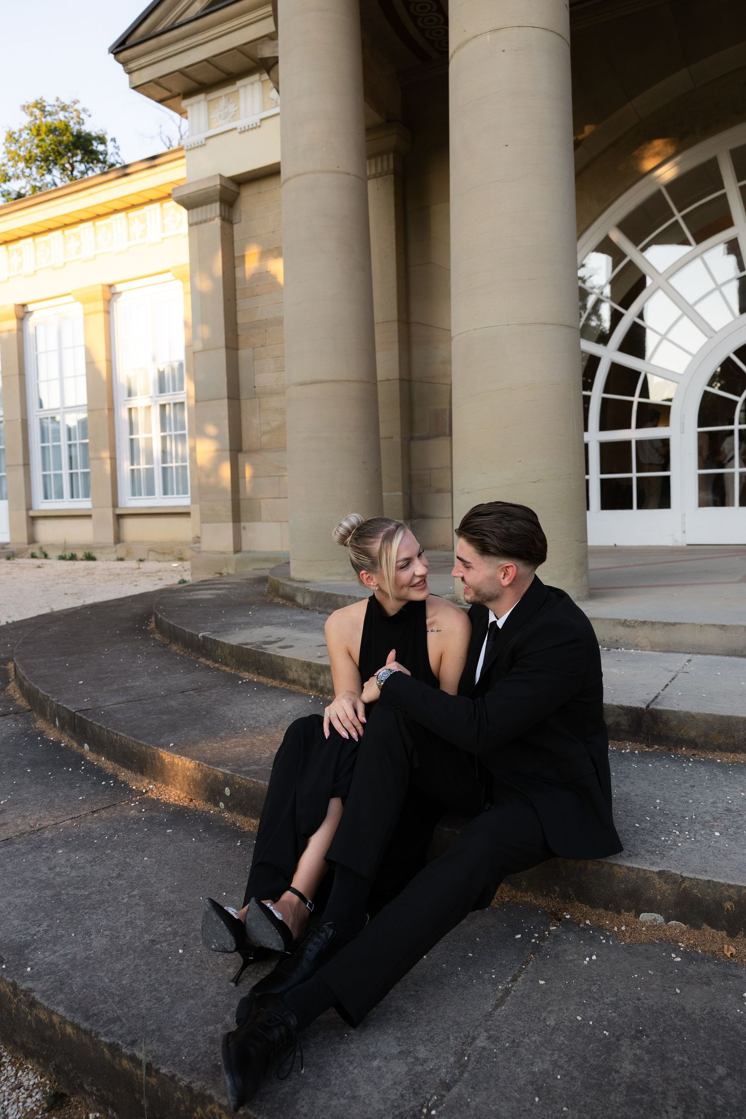 Couple in coordinating black formal outfits sit closely together on steps of a neoclassical building.