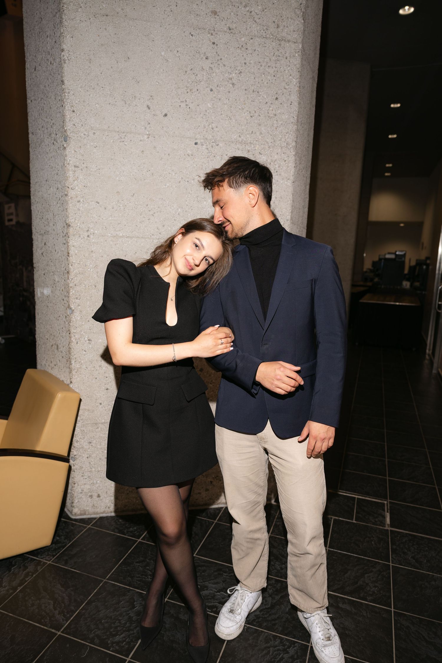 Couple in elegant attire share a romantic moment leaning against a concrete pillar at an indoor venue.