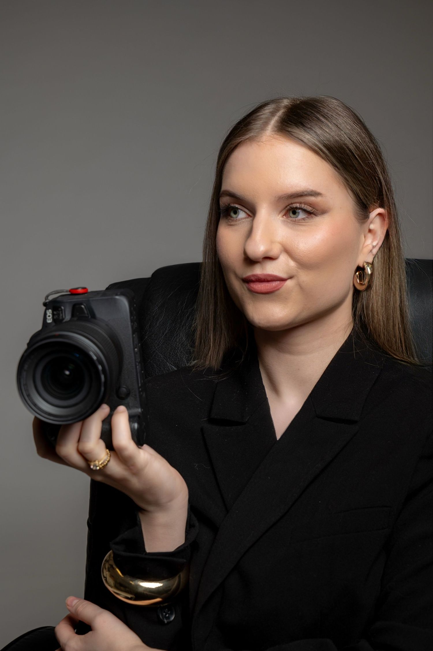 Young woman in black blazer holding a professional camera, wearing gold jewelry, posed against a gray background.