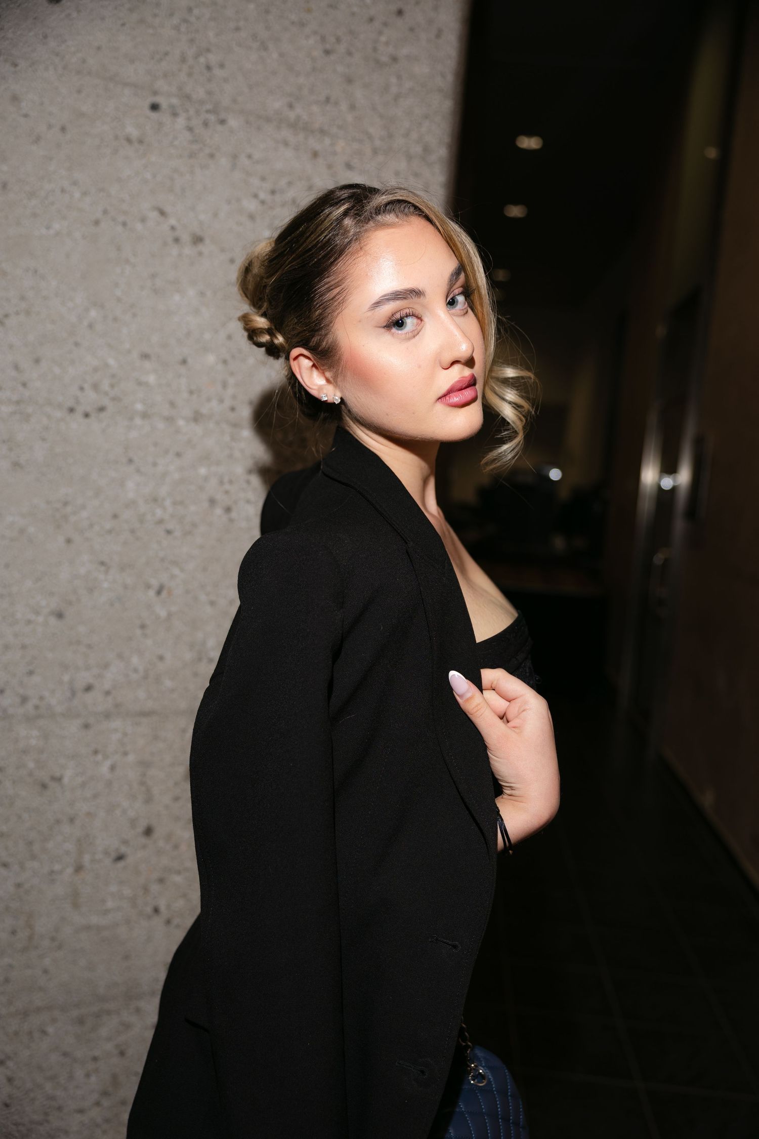 Young woman with elegant updo and red lips posing confidently in a black blazer in a dimly lit hallway.
