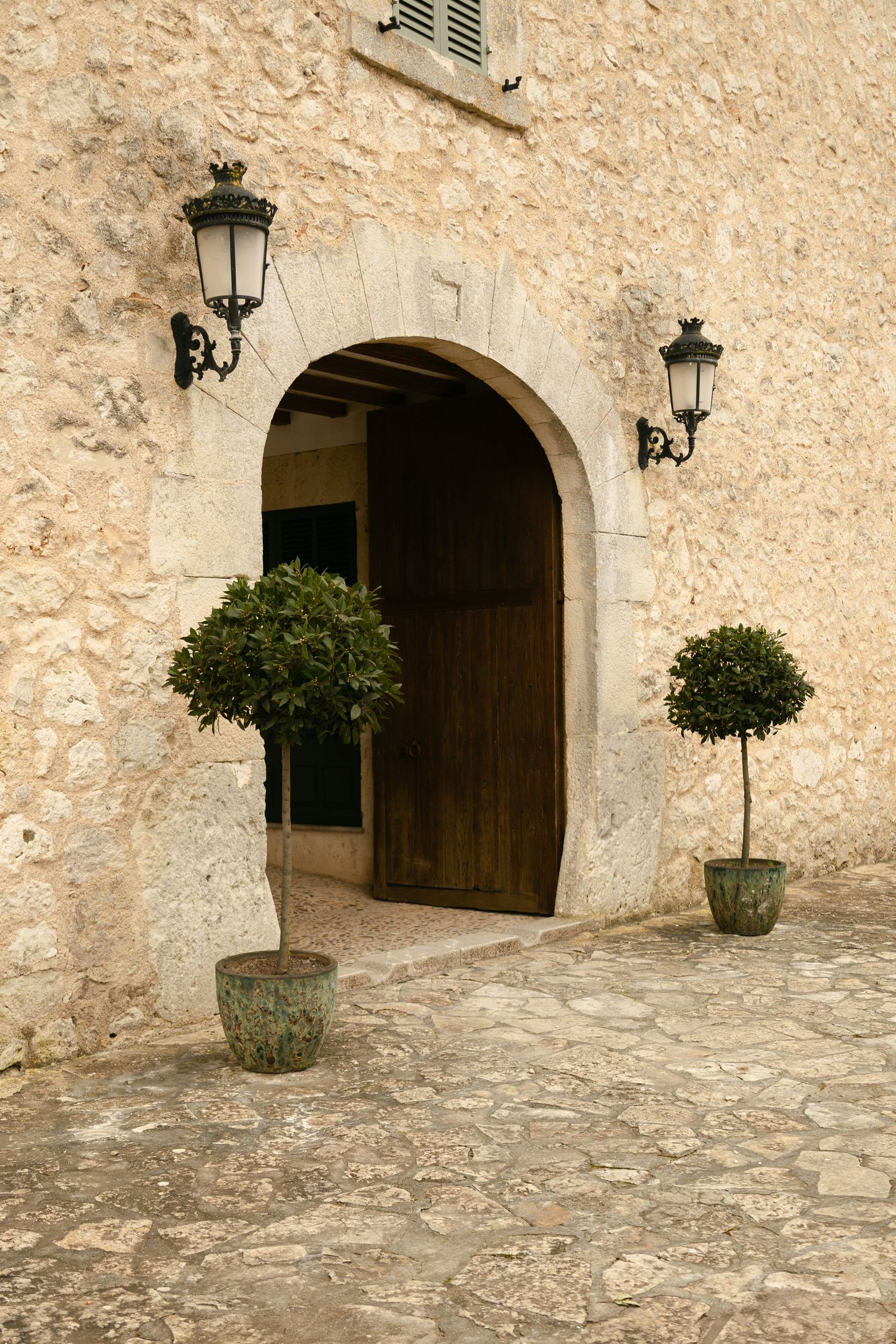 Stone archway entrance with wooden door, flanked by potted topiary trees and vintage wall lanterns.
