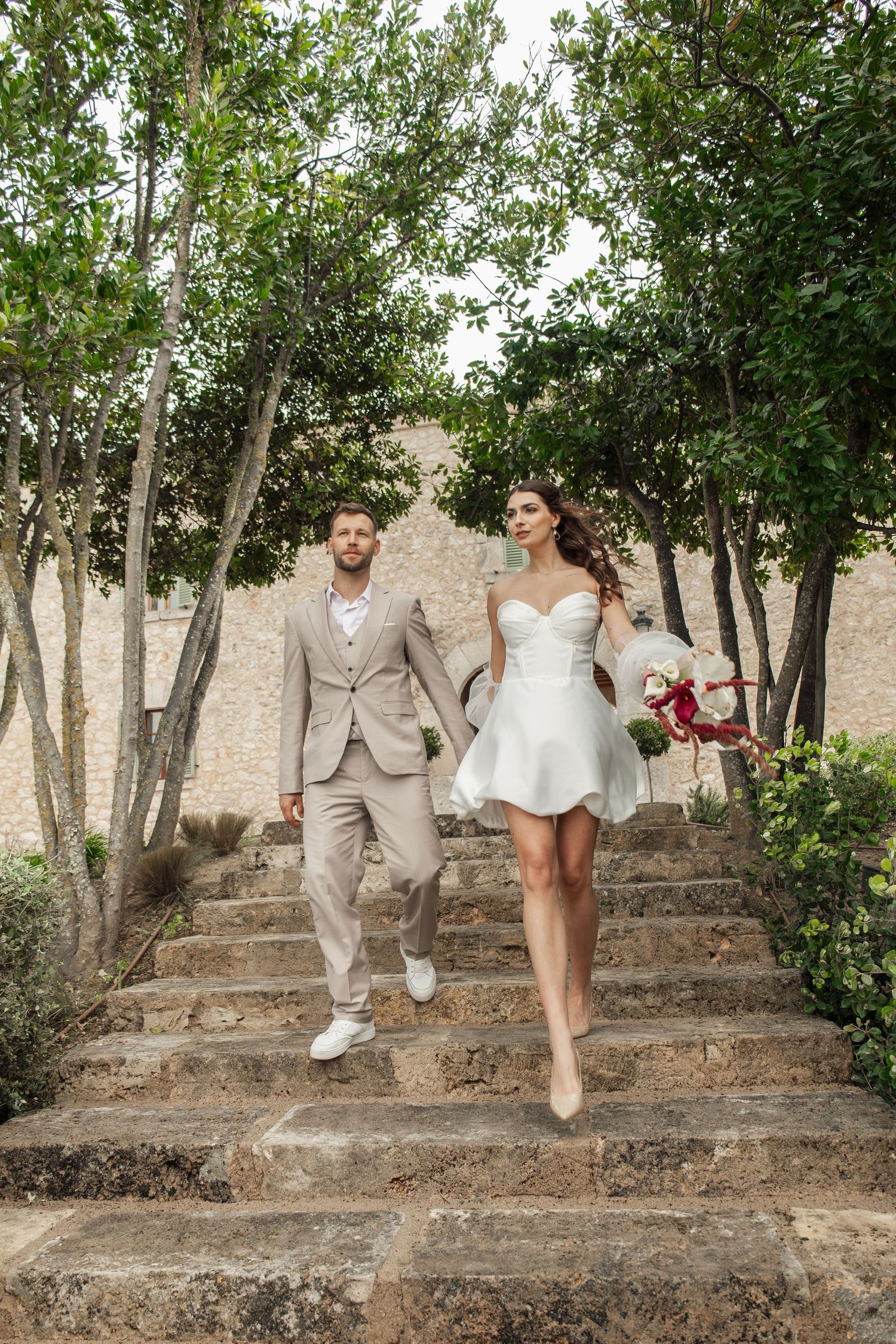 Bride in short white dress and groom in beige suit walking down stone steps surrounded by lush green trees.