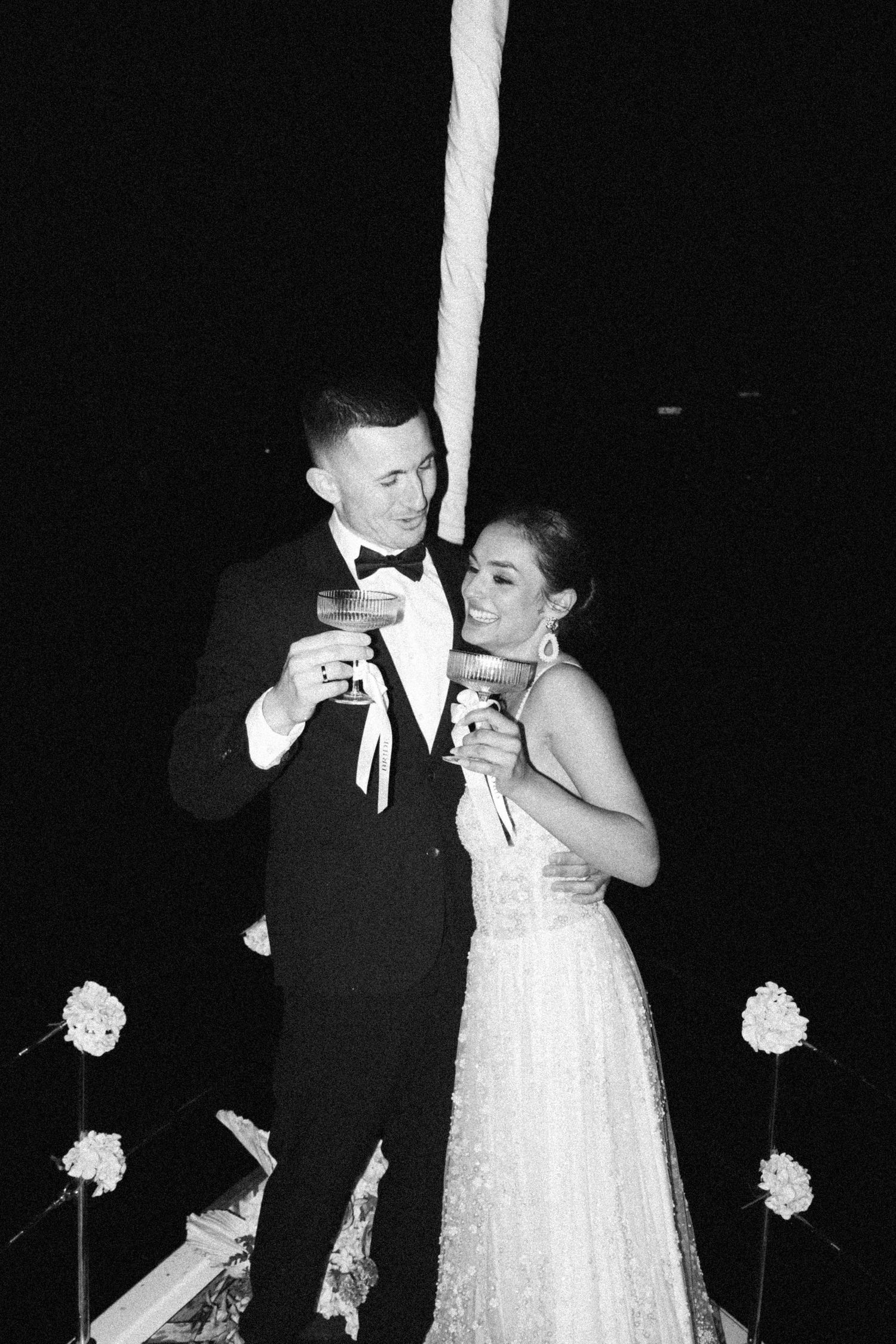 Black and white photo of bride and groom laughing and holding cocktails on boat at night.