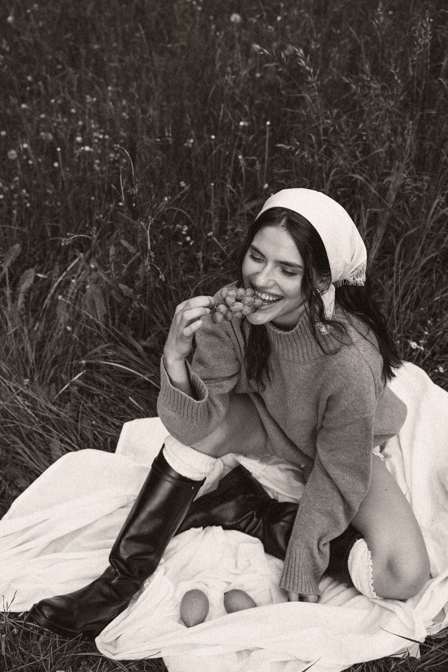 Black and white photo of smiling woman eating grapes in a wildflower field.