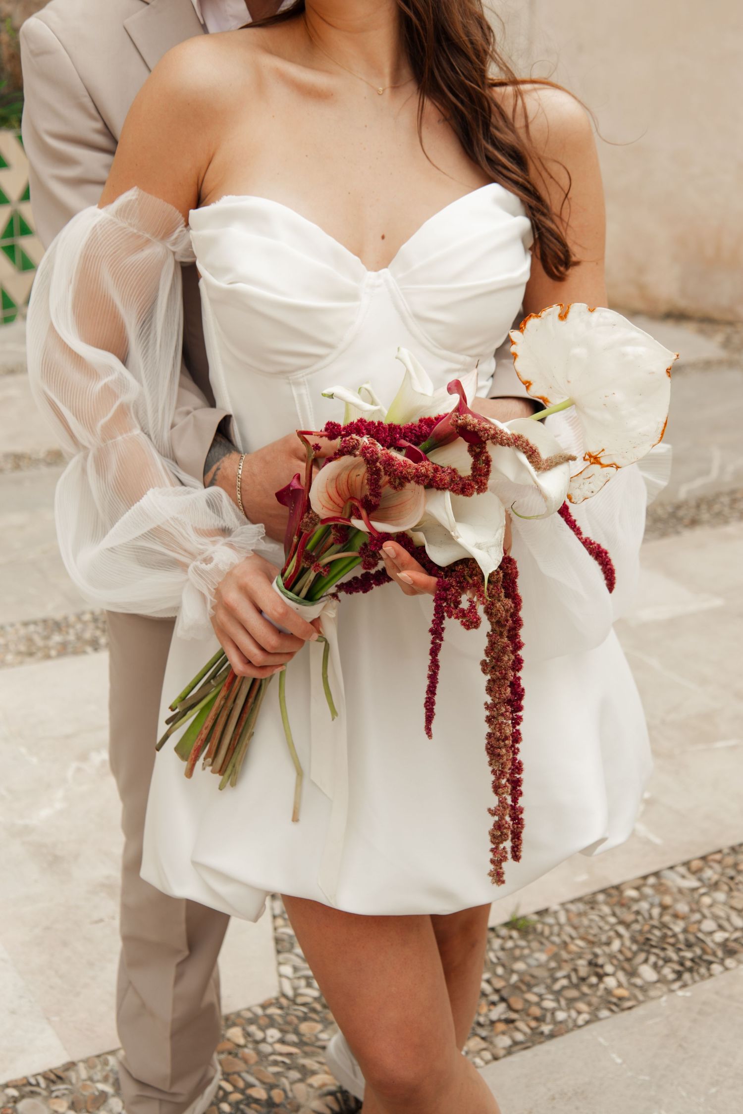 Bride in short white off-shoulder dress holding dramatic bouquet with red amaranthus and calla lilies.