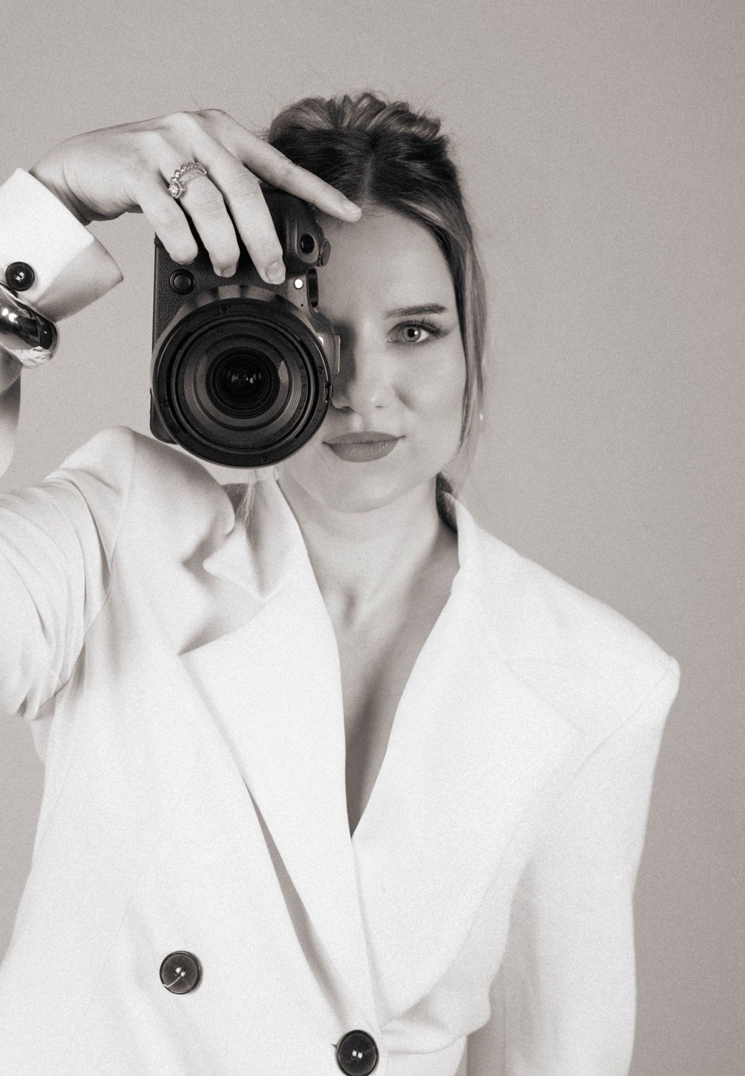 Black and white portrait of a woman in a white blazer holding a camera to her face.
