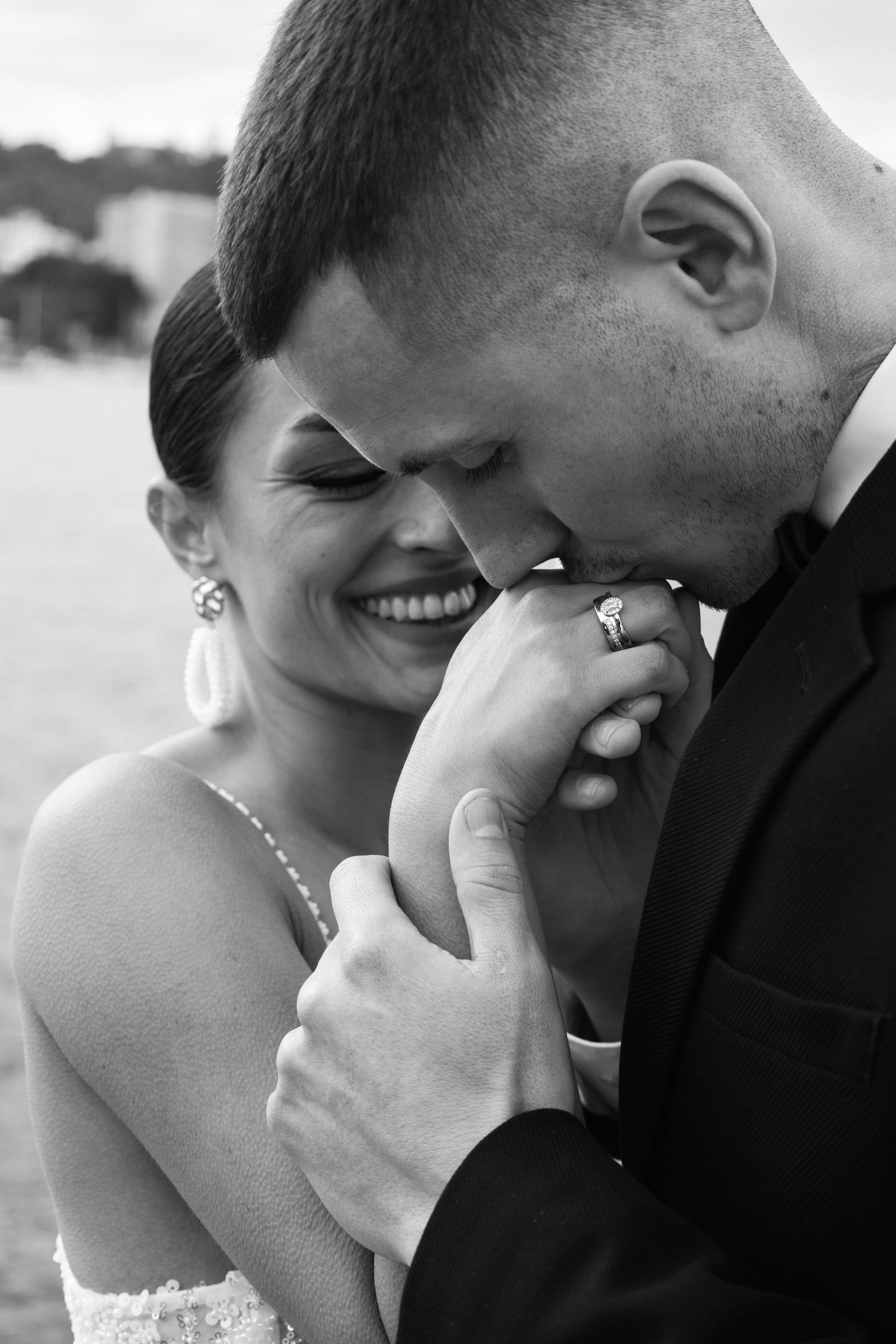 Groom tenderly kisses brides hand as she smiles wearing diamond ring, romantic black and white wedding portrait.