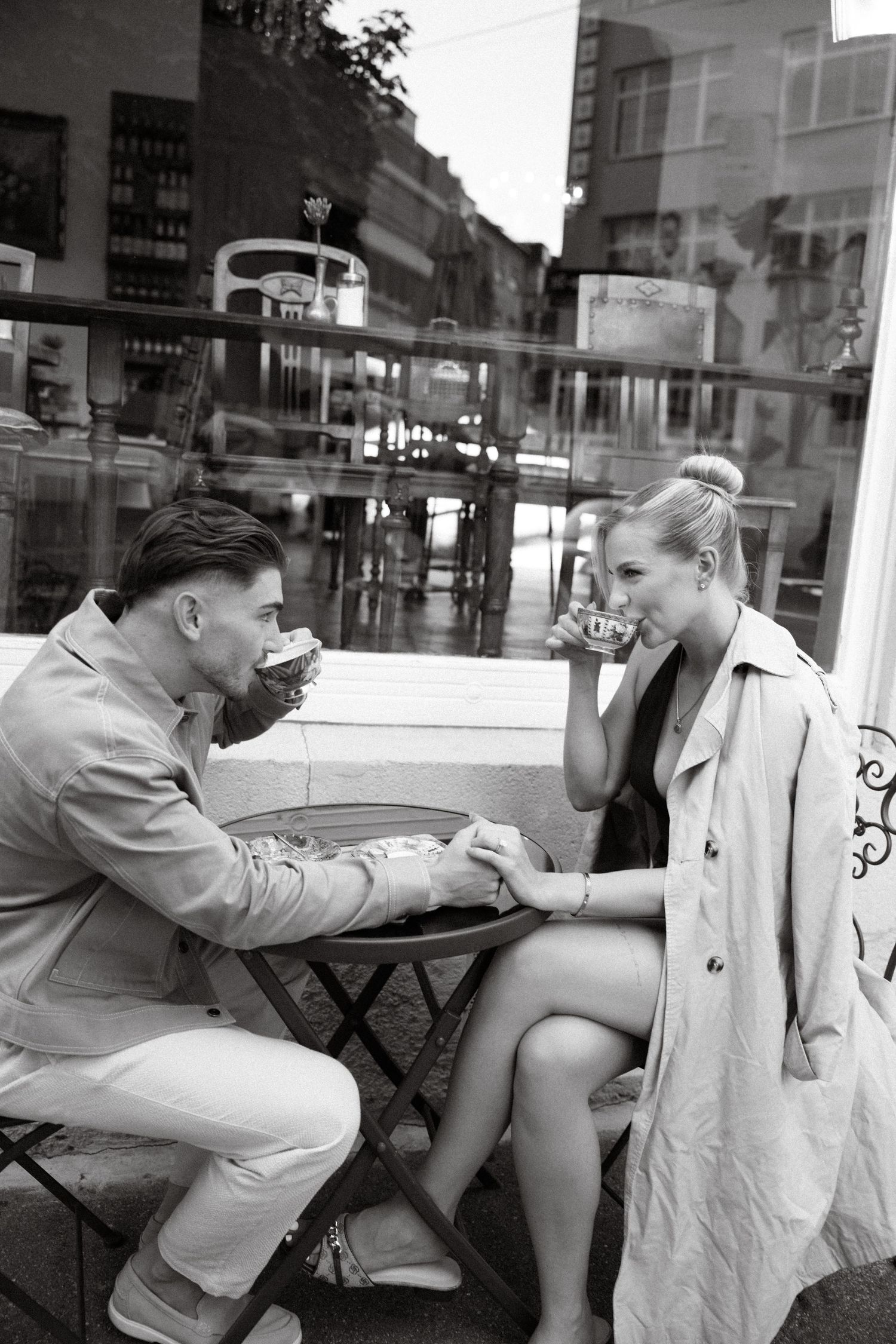 Black and white photo of a stylish couple holding hands and drinking coffee at a sidewalk cafe.