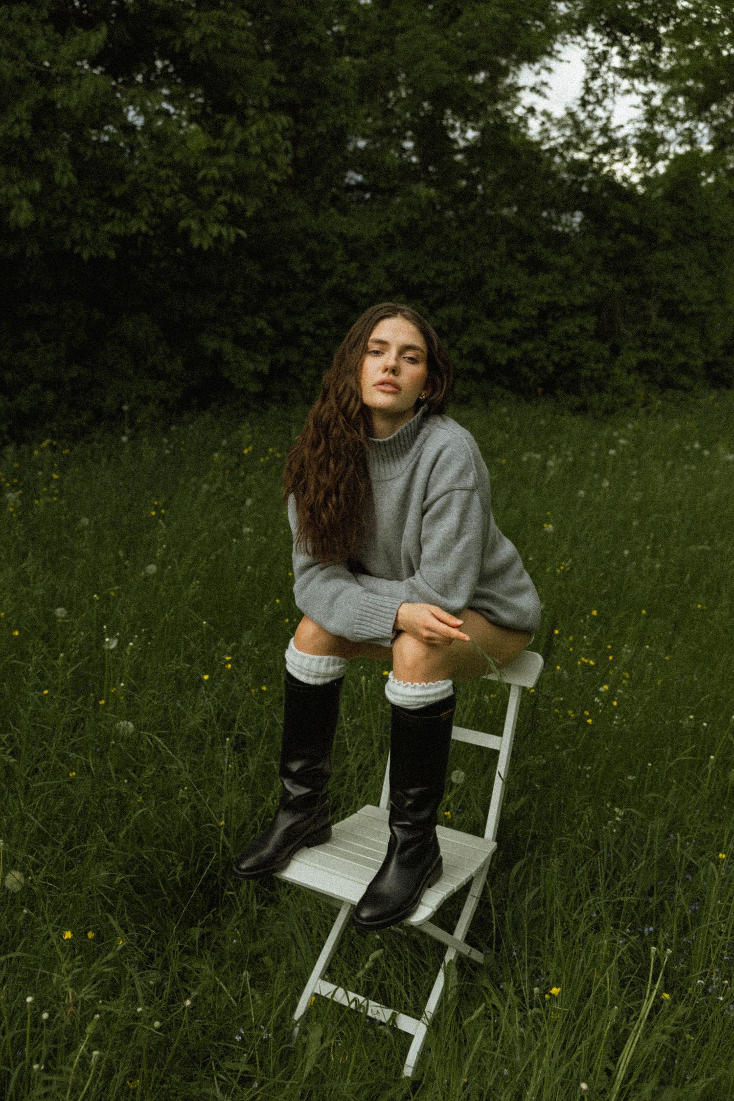 Woman in grey turtleneck sweater and black boots sitting on white chair in flower field.