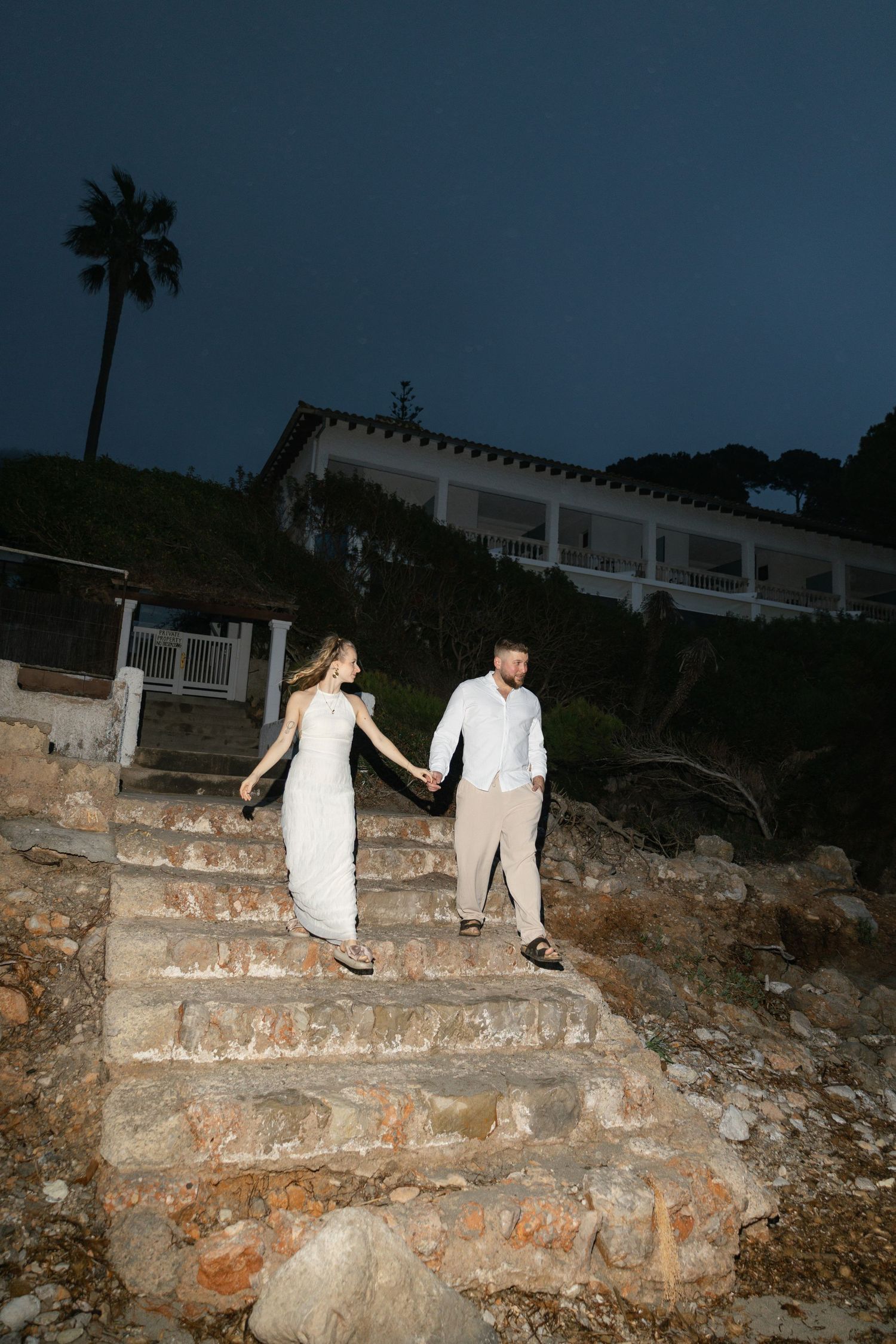 Bride and groom holding hands walking down stone steps at night with a white villa and palm tree in the background.