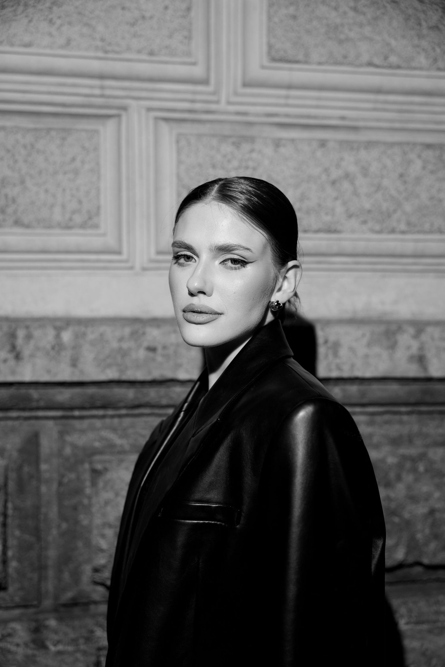 Black and white portrait of a young woman in a leather jacket posing against a stone wall.