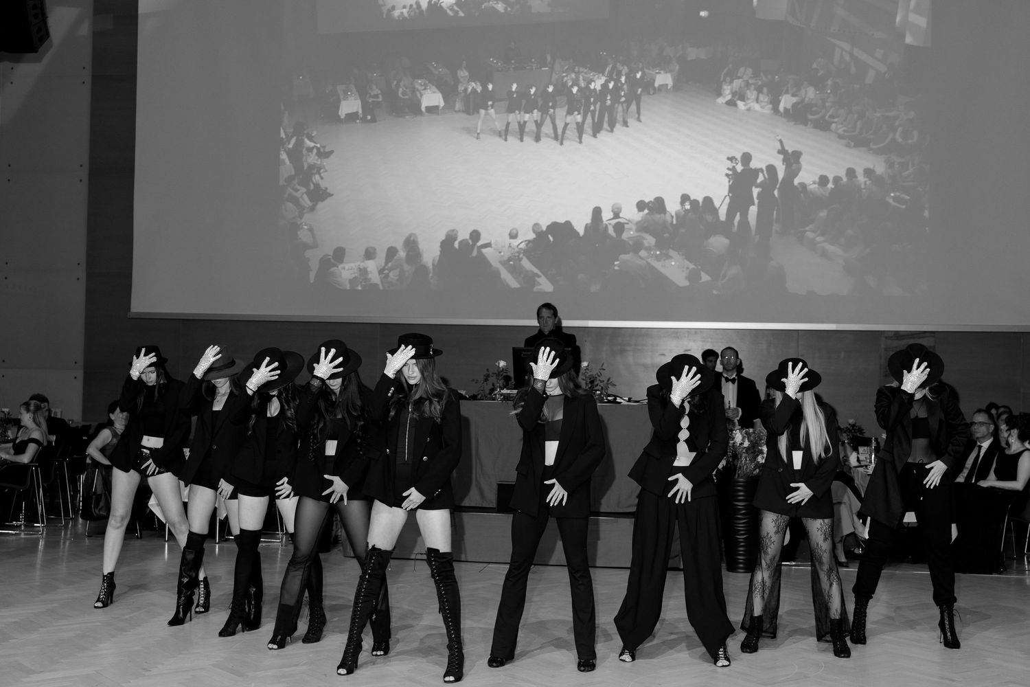 Black and white photo of female dance troupe in black outfits performing synchronized routine at gala event.