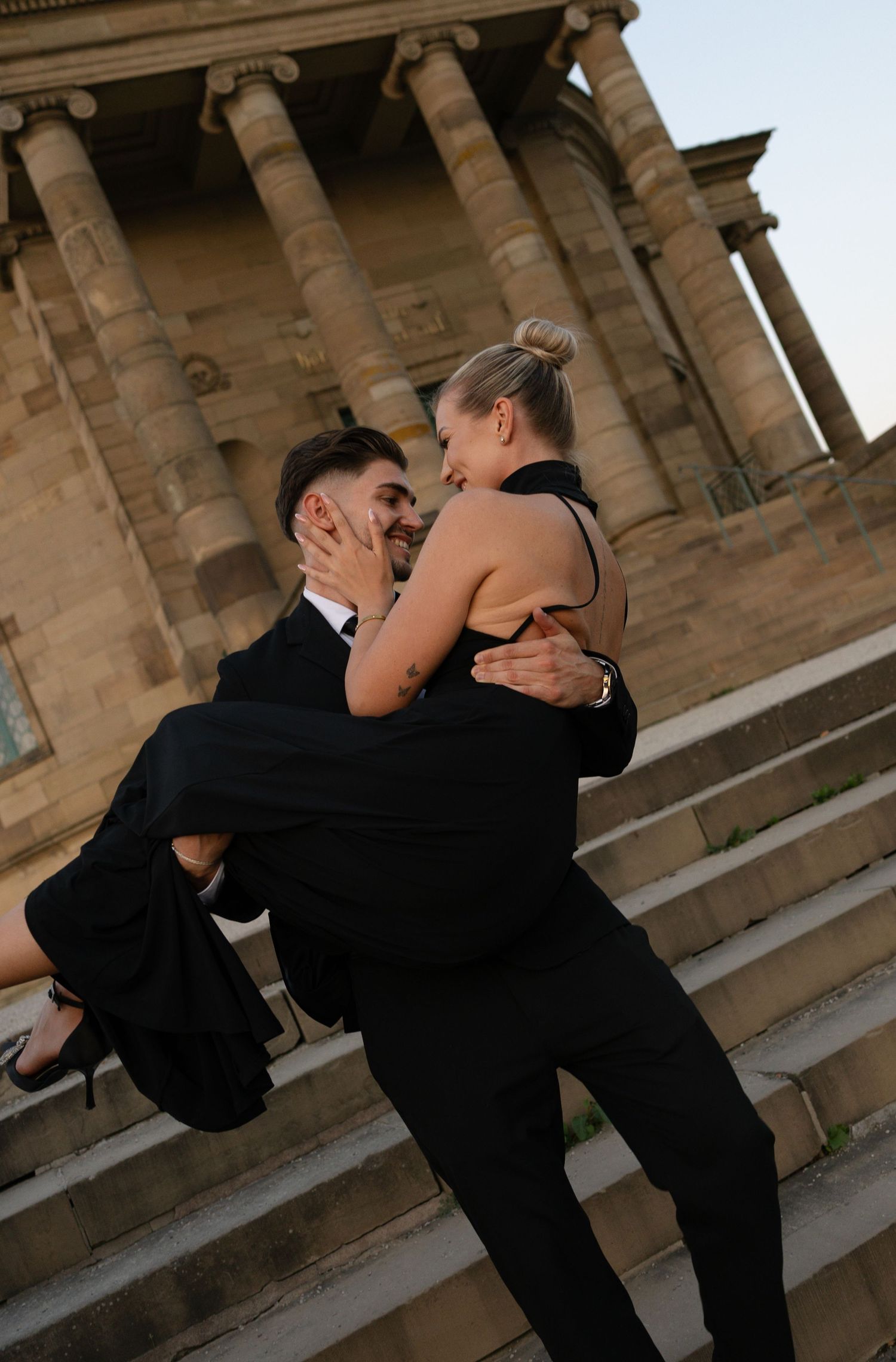A man in a black suit lifts a woman in a black dress on stone steps before a classical columned building.