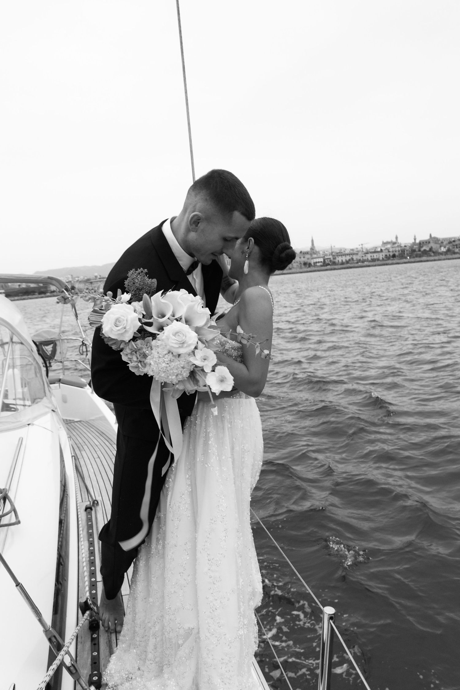 Black and white photo of bride and groom kissing on sailboat deck holding white floral bouquet.