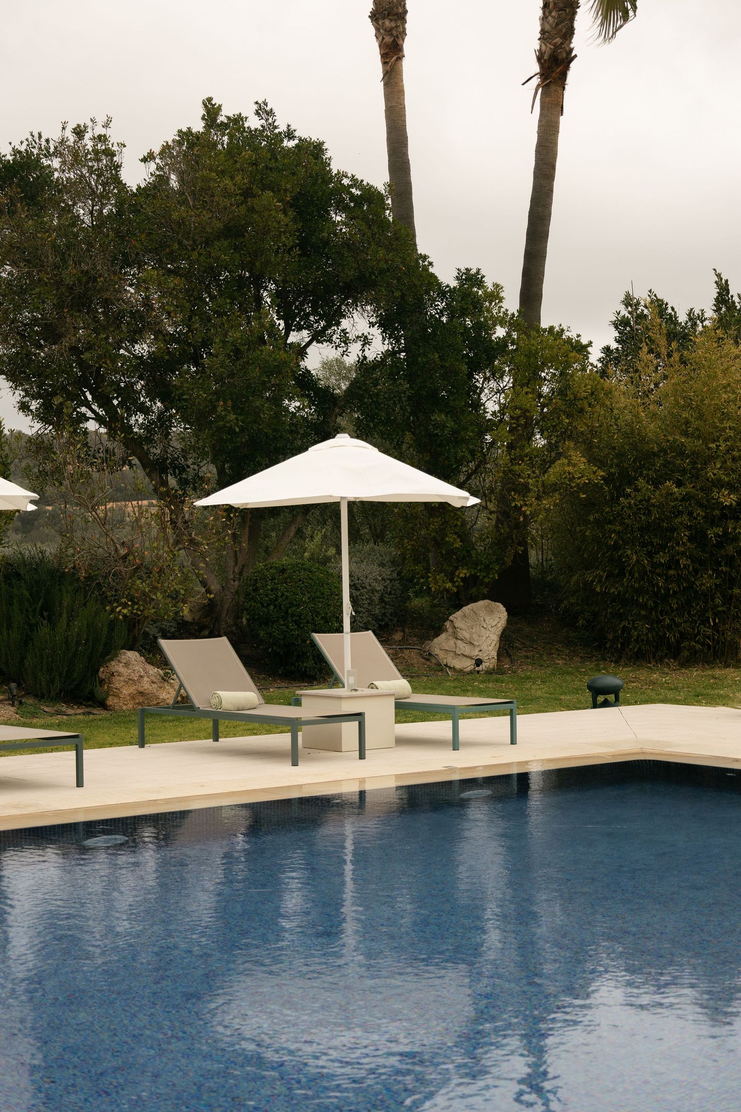 Poolside lounge chairs with white umbrella beside a calm blue swimming pool surrounded by palm trees.