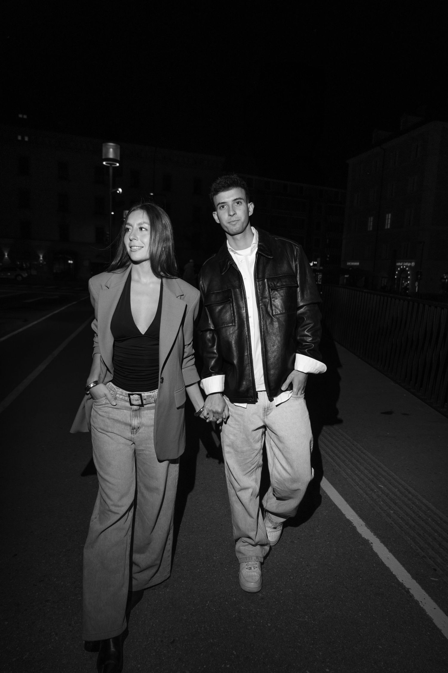Black and white photo of a couple holding hands while walking on a city street at night.