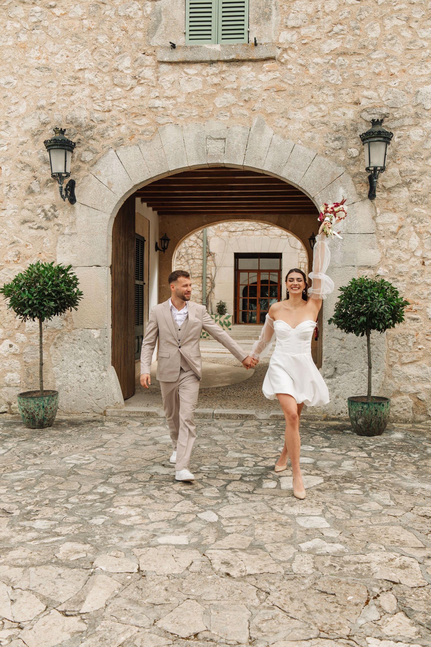 Bride and groom holding hands walking through stone archway at rustic European venue on wedding day.