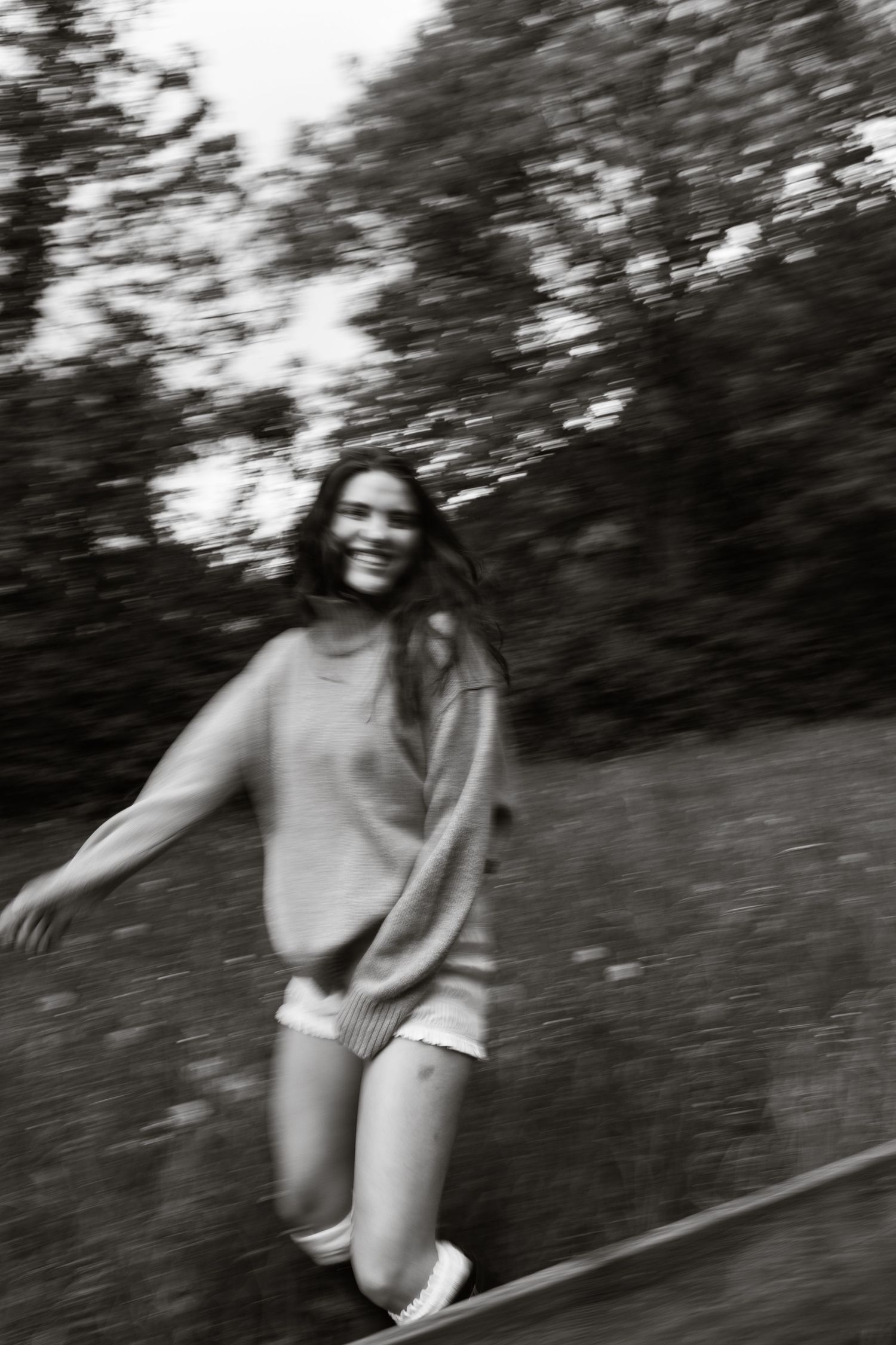 Black and white motion blur photo of joyful woman running and smiling outdoors among trees.