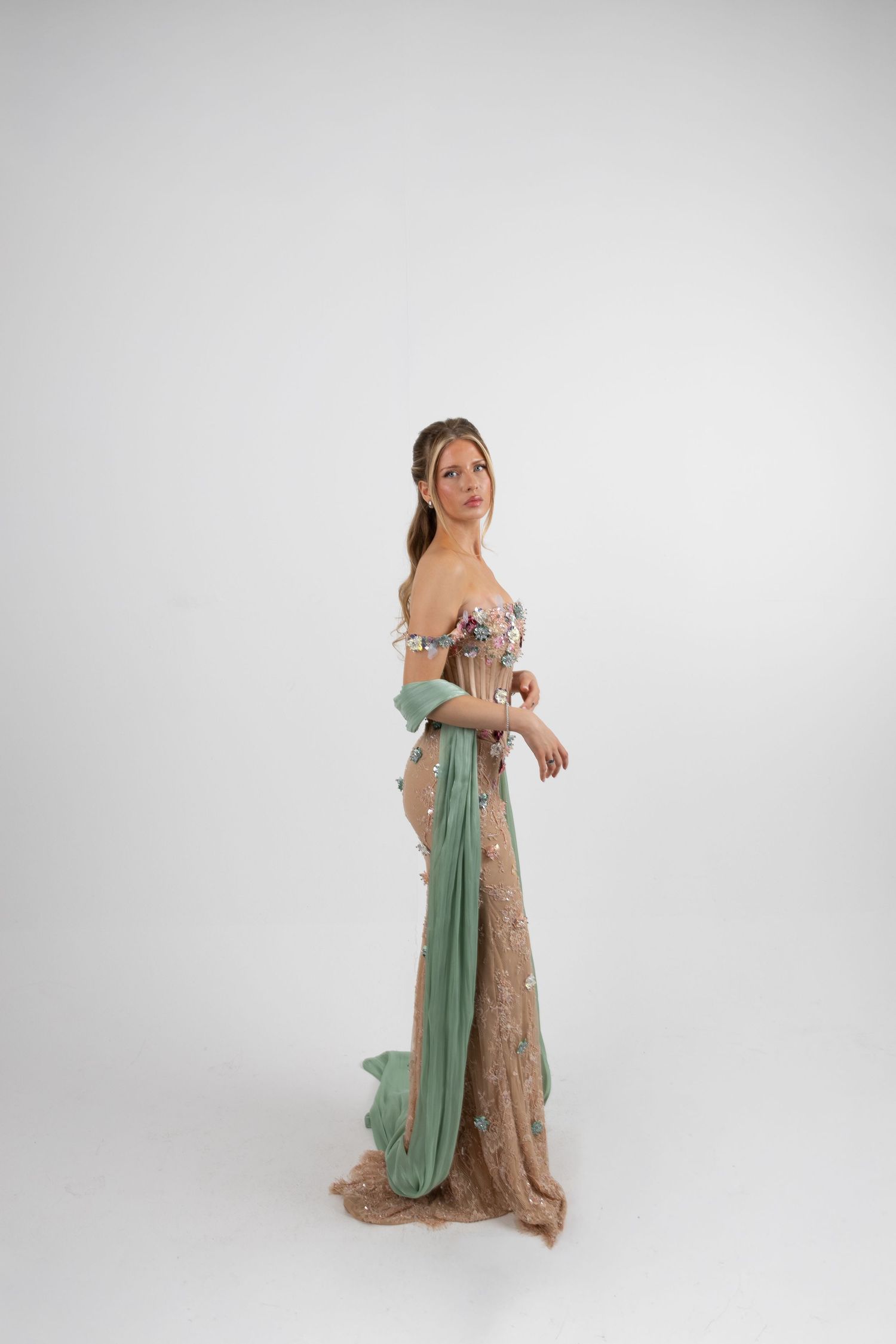 Woman in elegant sage green and floral off-shoulder gown with delicate embellishments poses gracefully on white background.