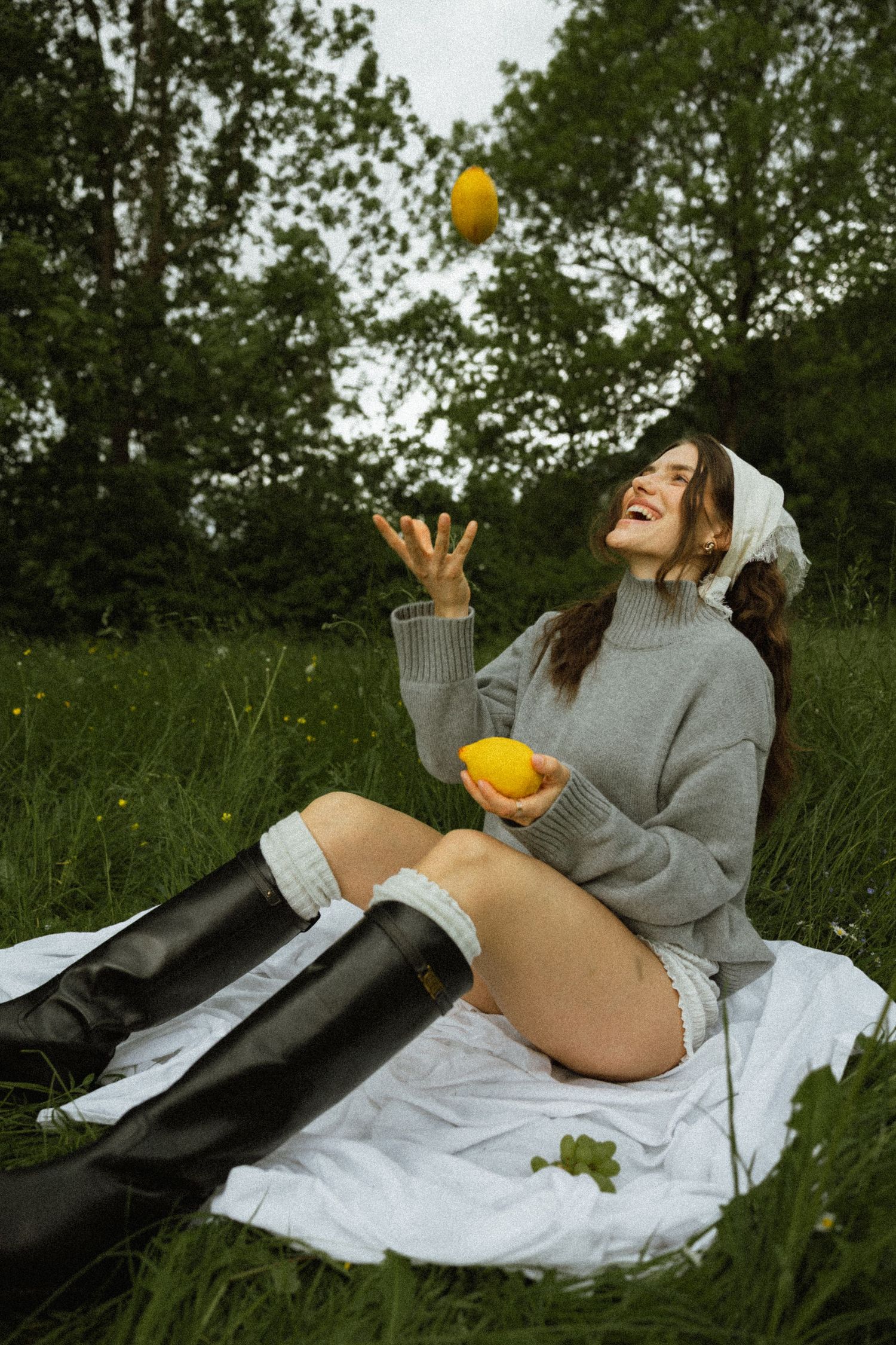 Laughing woman in gray sweater and black boots tossing lemons while sitting on blanket in green meadow.