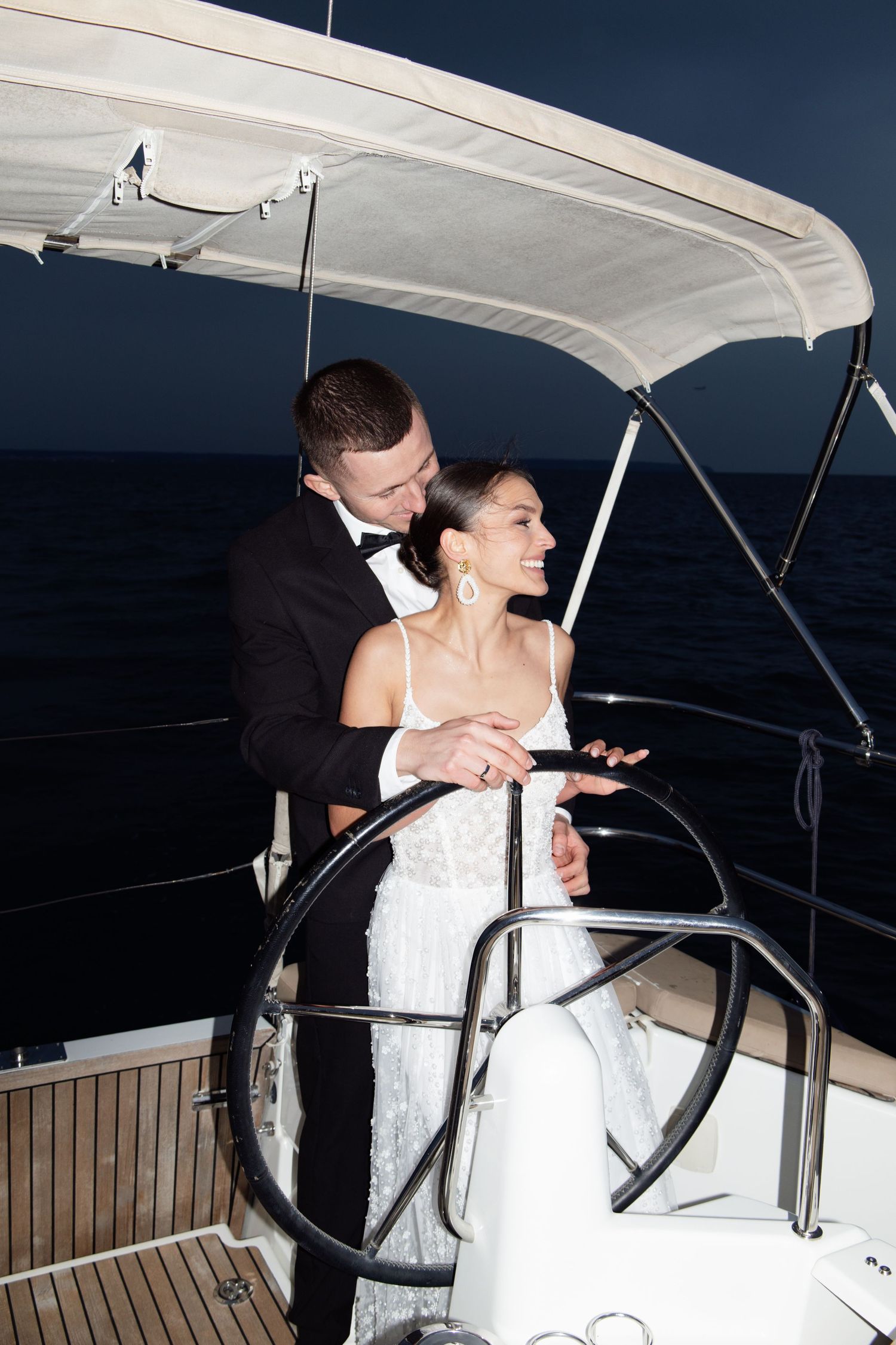 Elegant bride and groom in formal attire steering a yacht together at night on the open ocean.