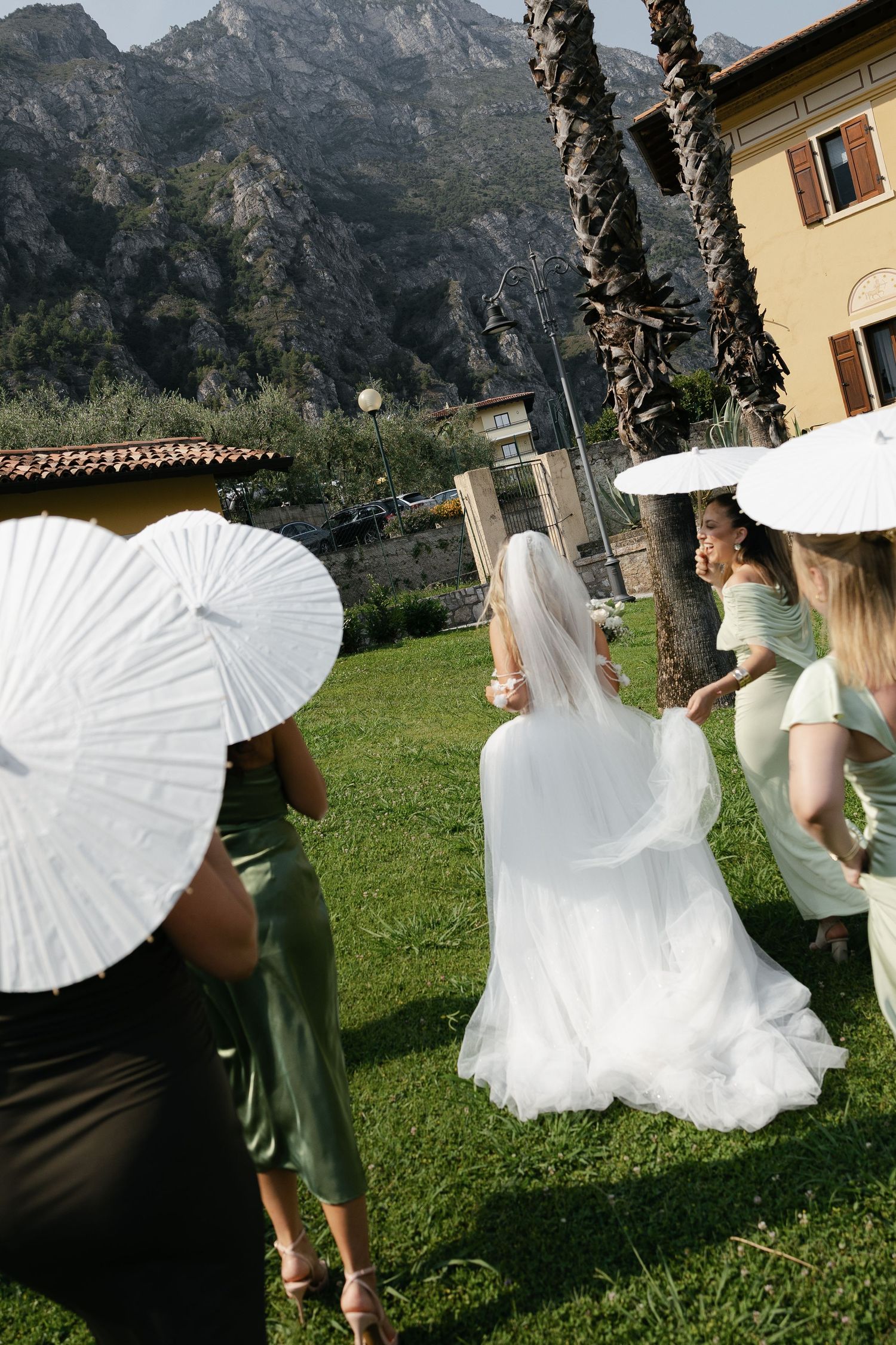 Bride in white gown with bridesmaids holding parasols in a garden with mountain backdrop.