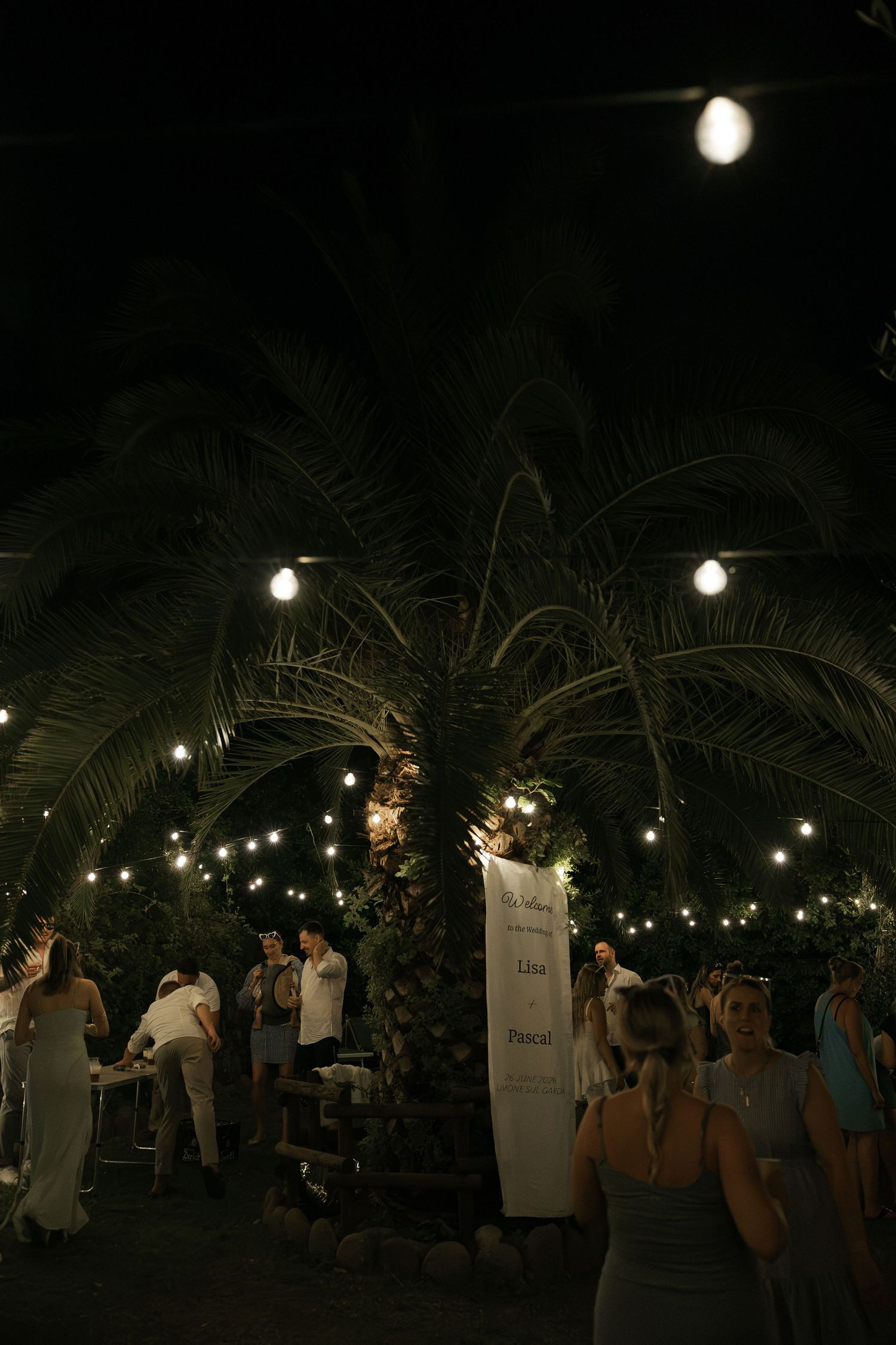 Outdoor night wedding reception under palm trees with string lights and guests dressed in white attire.