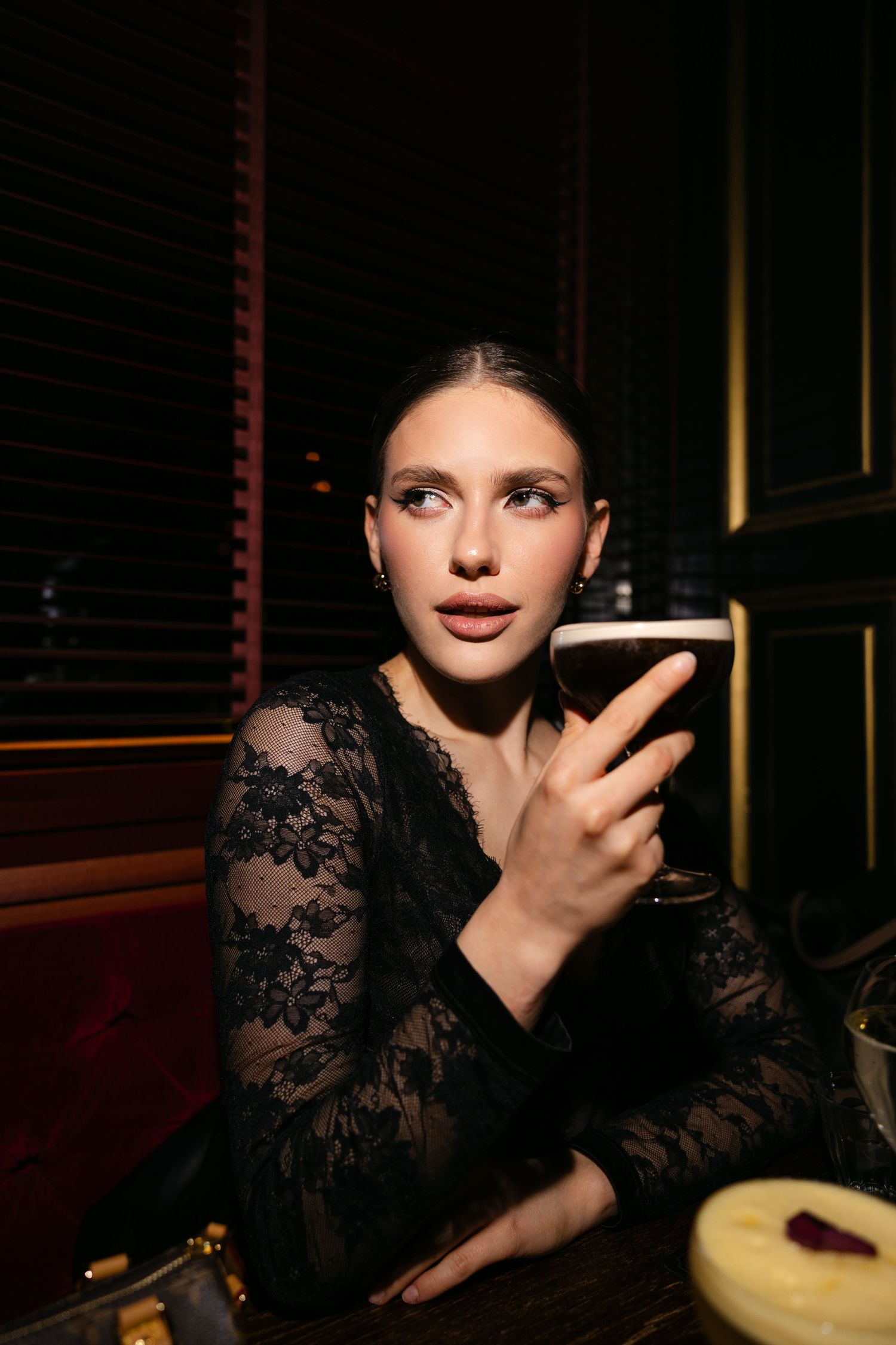 Young woman in black lace dress holding dark beer glass in dimly lit bar.
