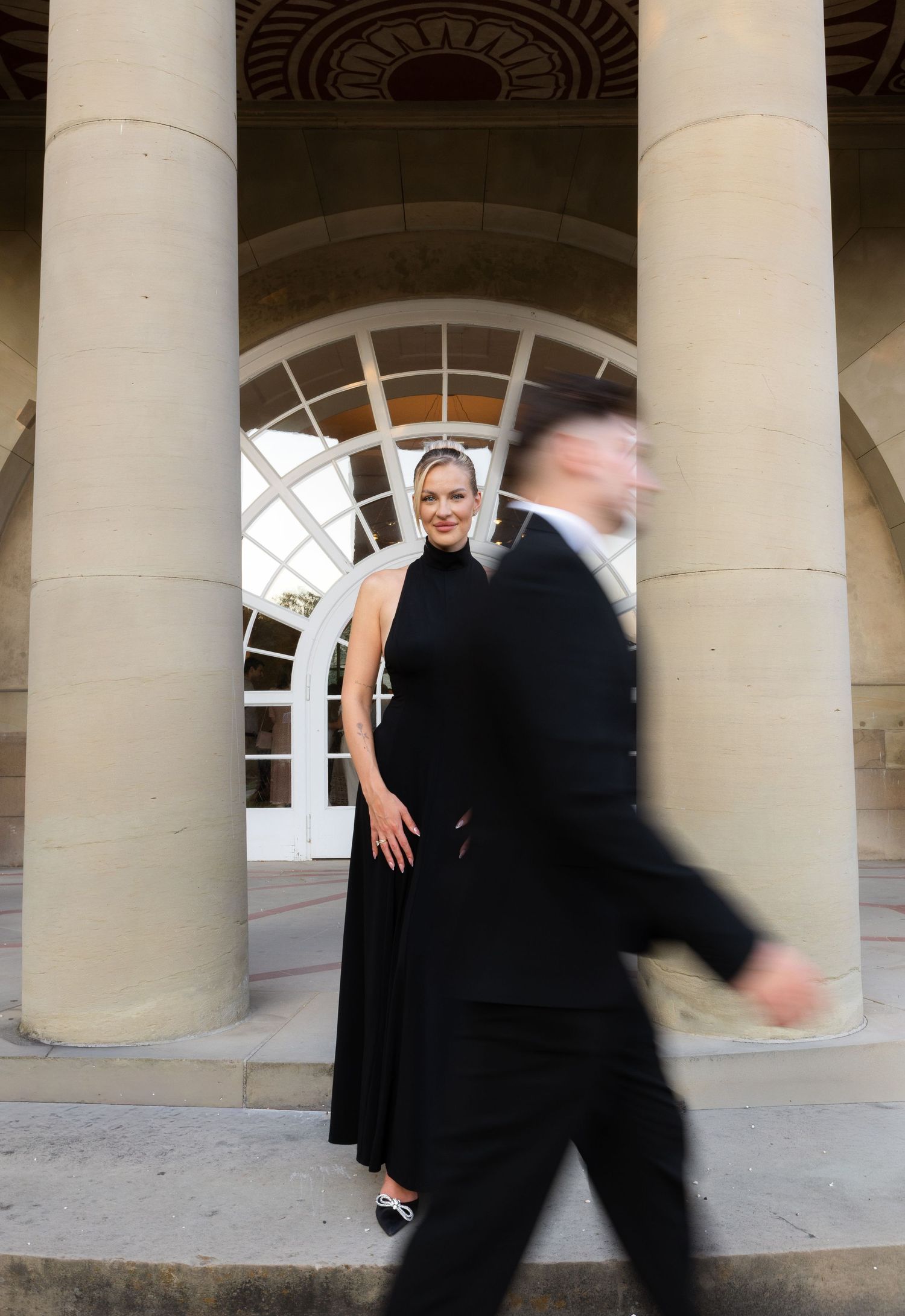 Elegant woman in black halter dress poses between classical columns as man walks past in blur.