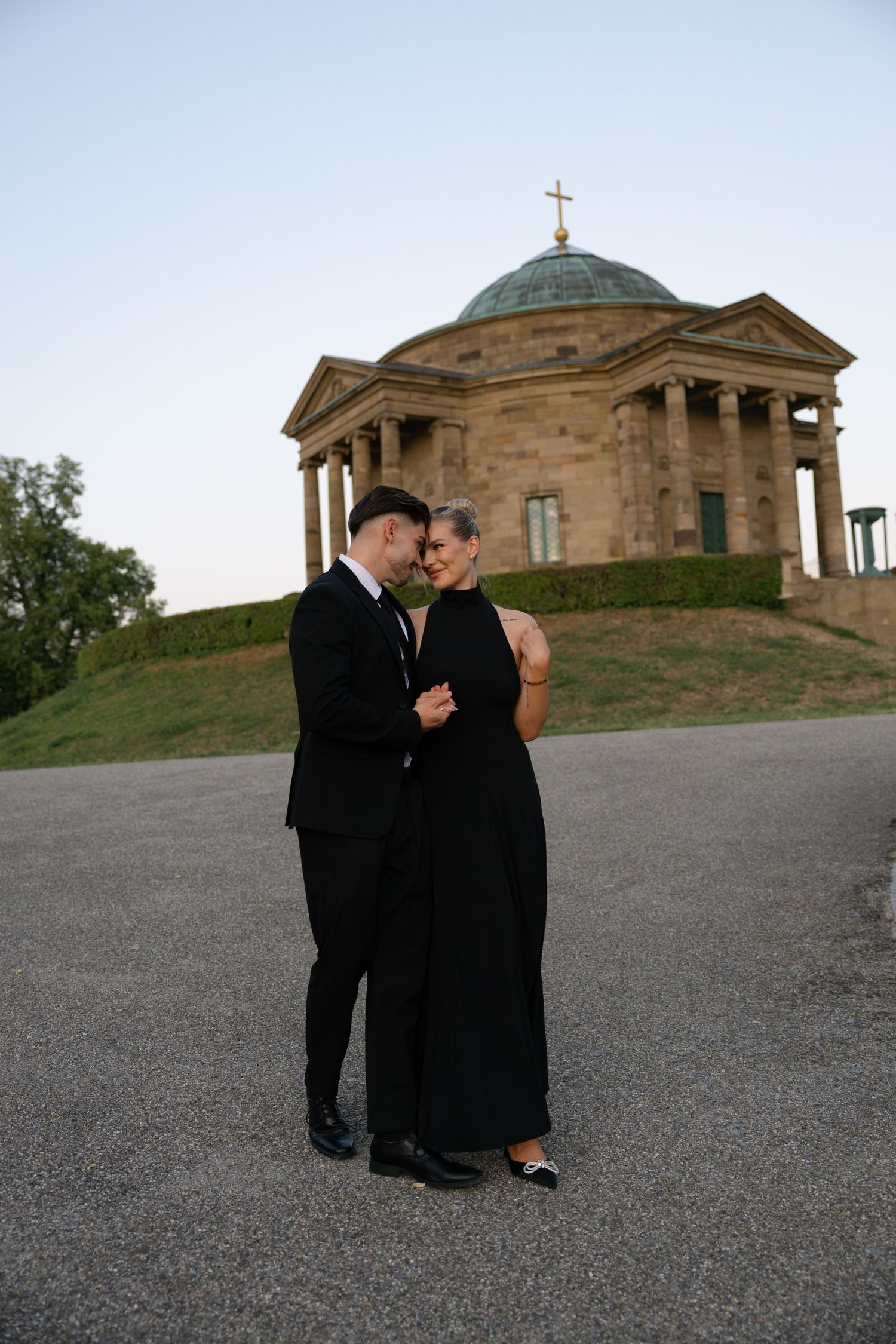 Couple in black formal attire embracing in front of a neoclassical domed chapel with a cross at dusk.