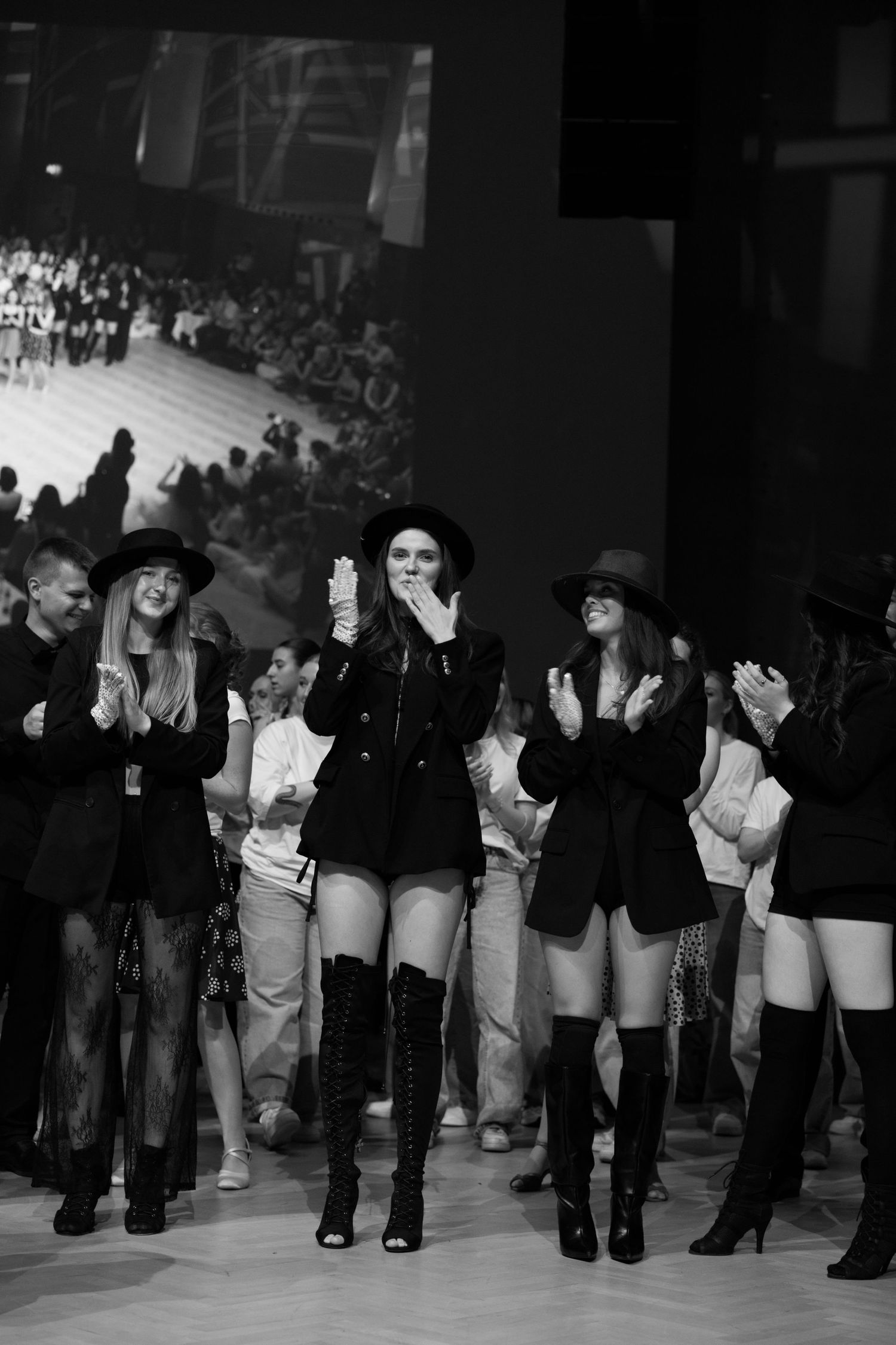 Black and white photo of models in black outfits and hats taking a bow on a fashion show runway.