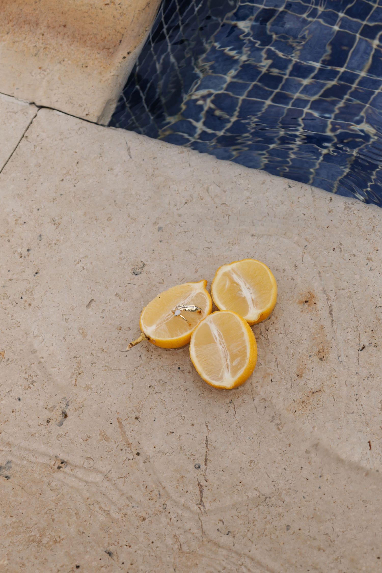 Three halved lemons resting on a stone pool deck beside a blue tiled swimming pool.