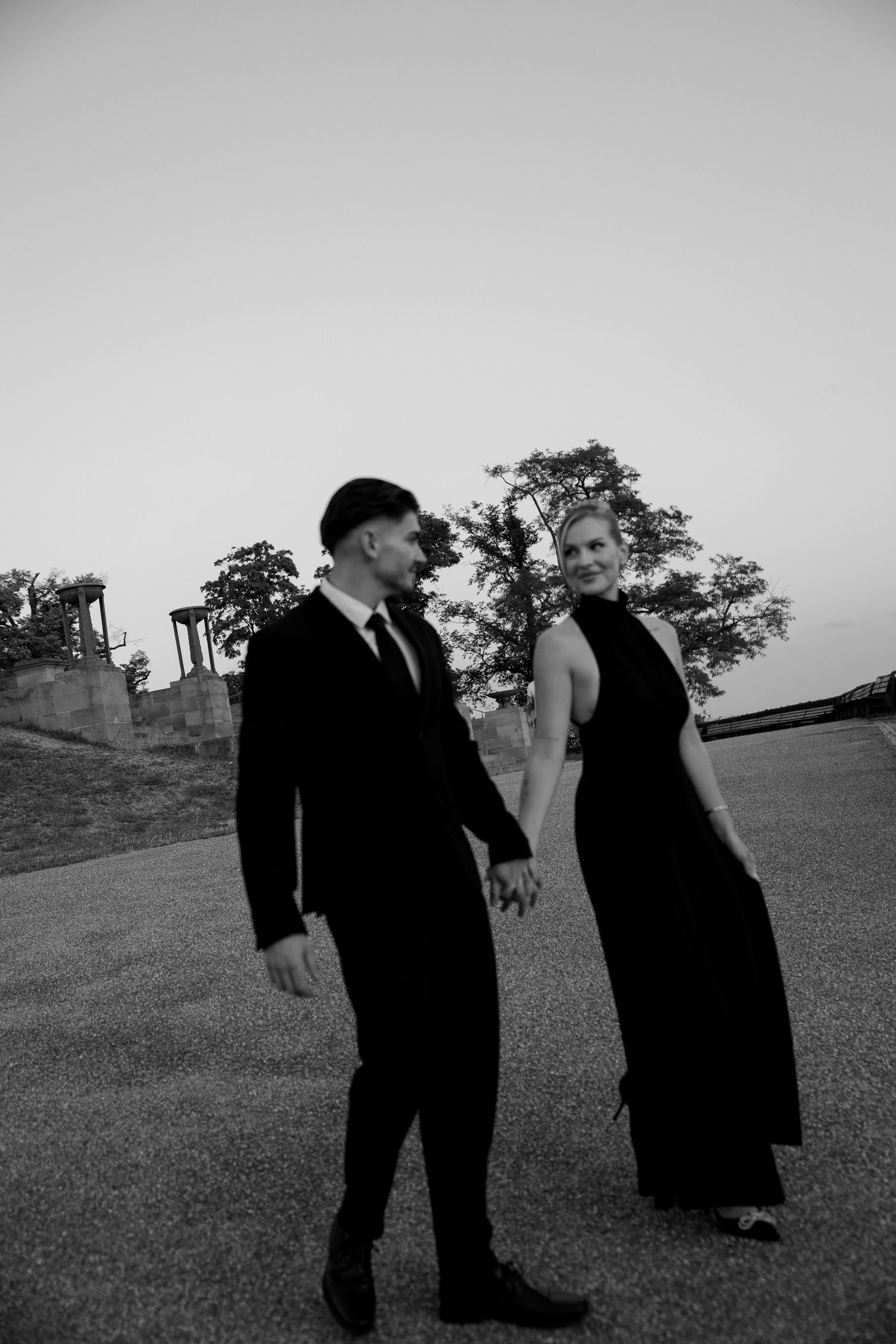 Black and white photo of an elegant couple holding hands, man in suit and woman in long gown, walking outdoors at dusk.