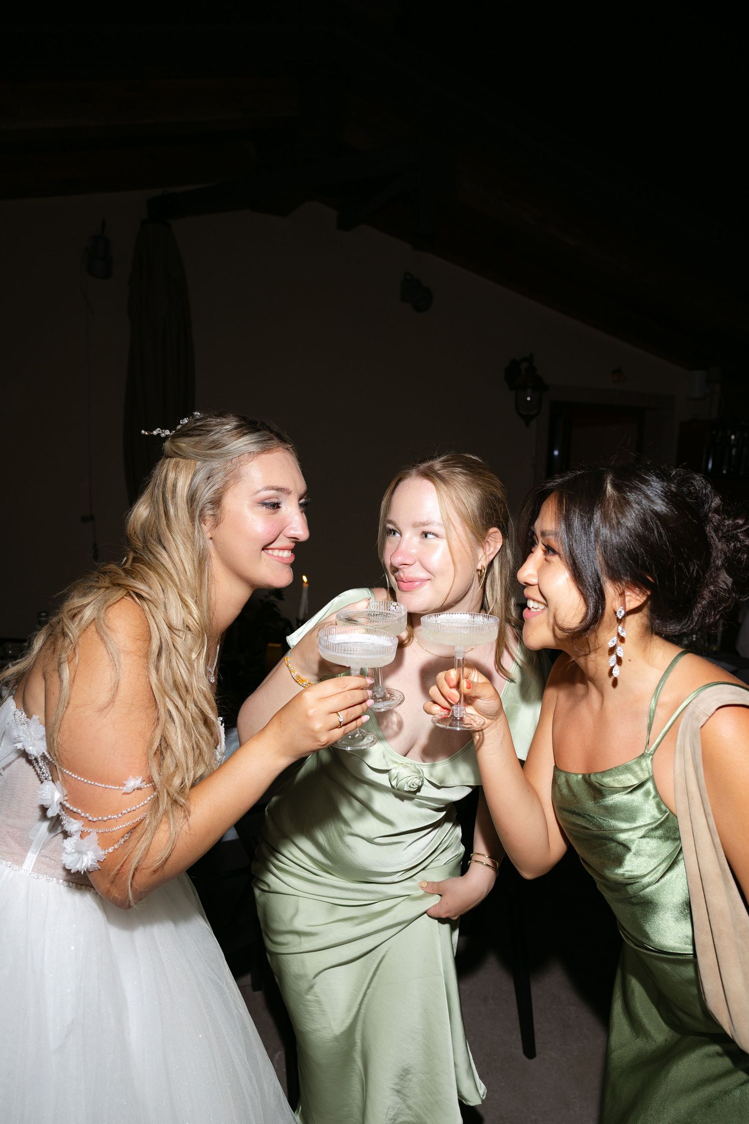 Bride in white gown toasting with two bridesmaids in sage green dresses at a wedding reception.