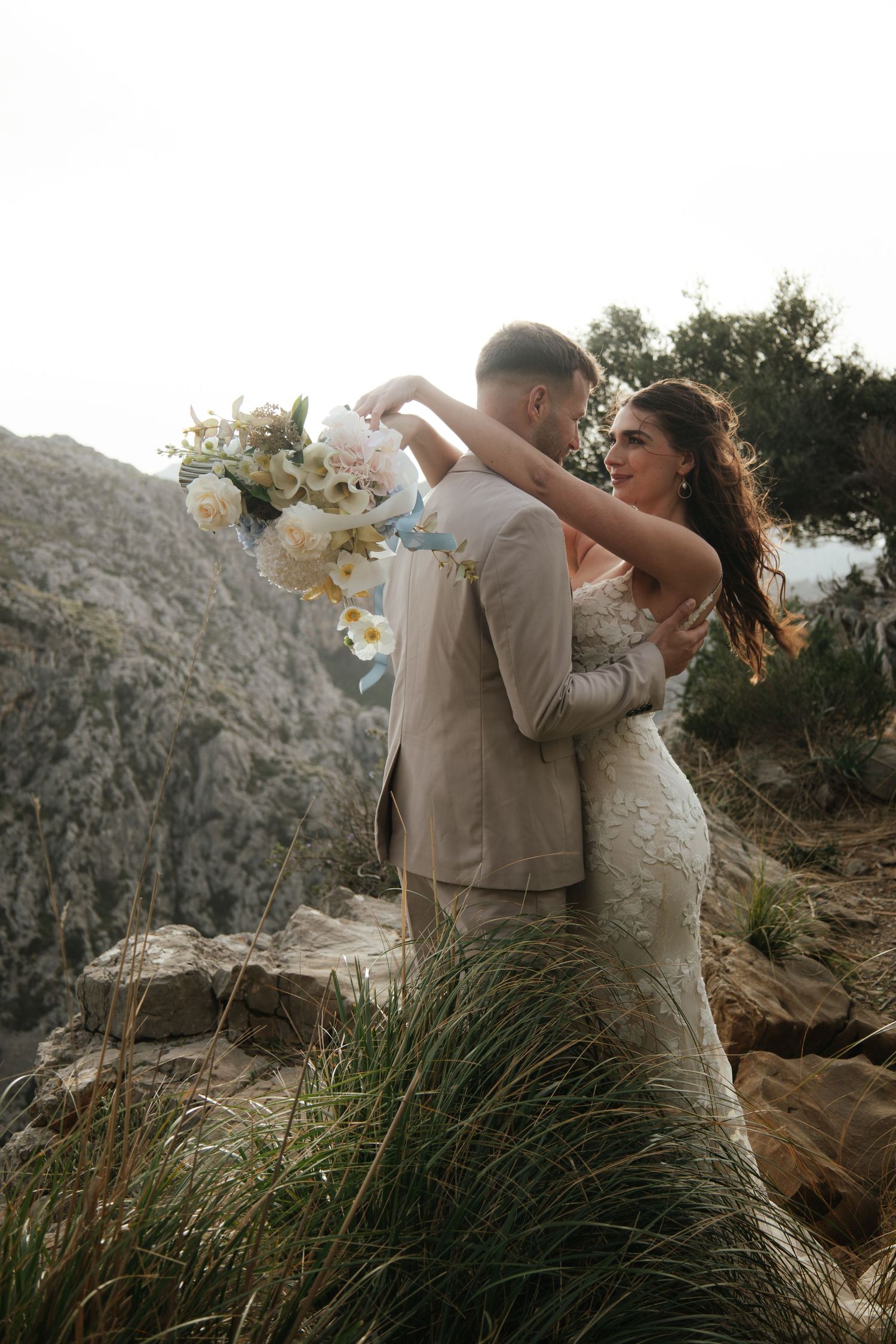 Bride and groom embrace on rocky cliffside, she holds white floral bouquet, wearing lace gown, he in beige suit.