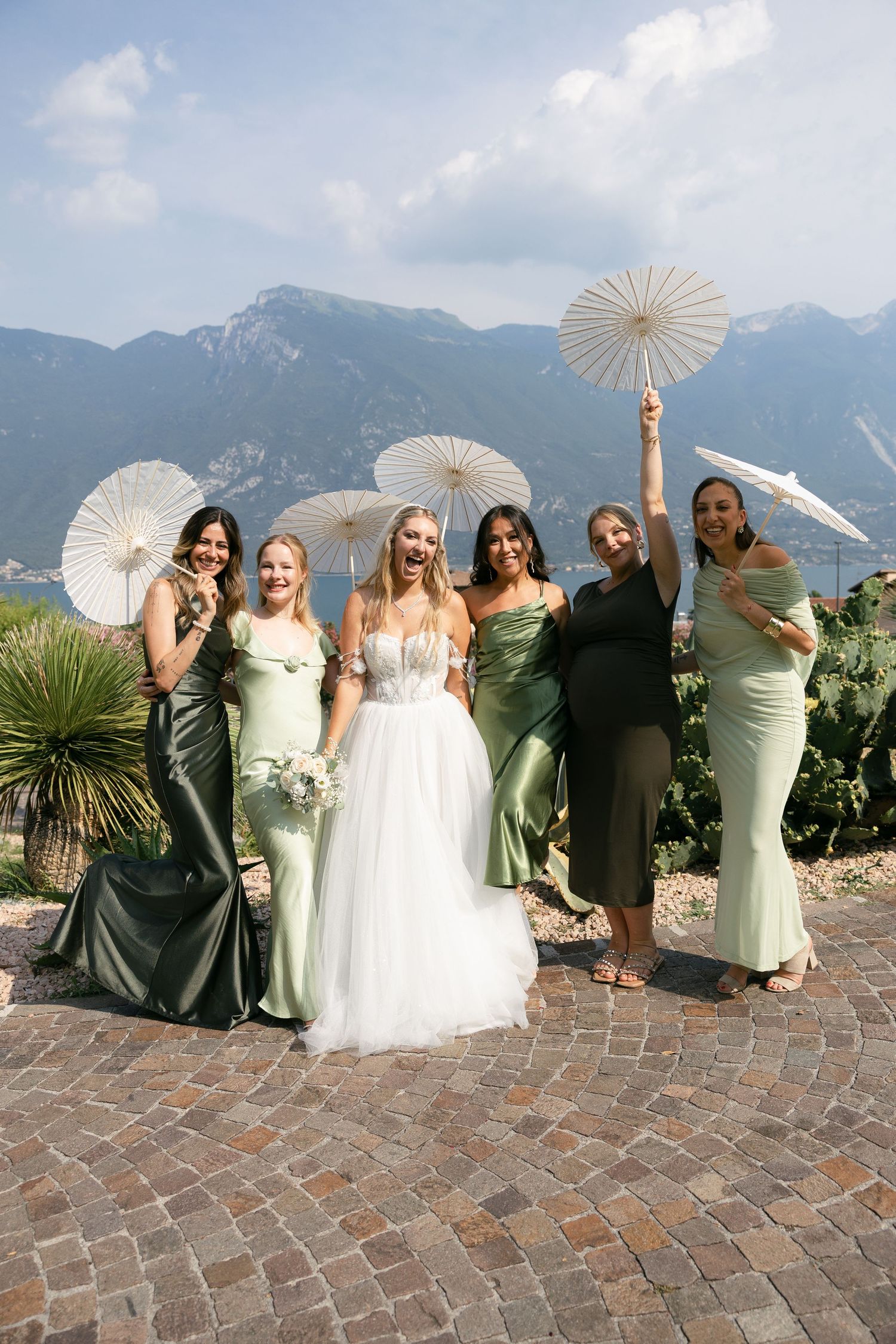 Bride with bridesmaids in sage green gowns holding parasols posing with stunning mountain lake backdrop.