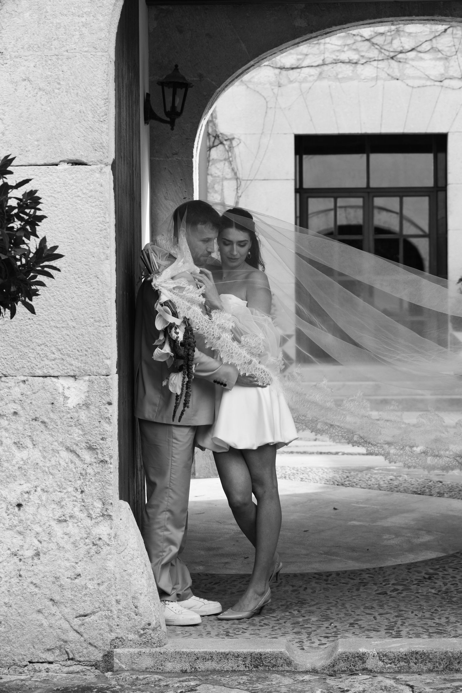 Black and white photo of bride and groom embracing under stone arch with veil.