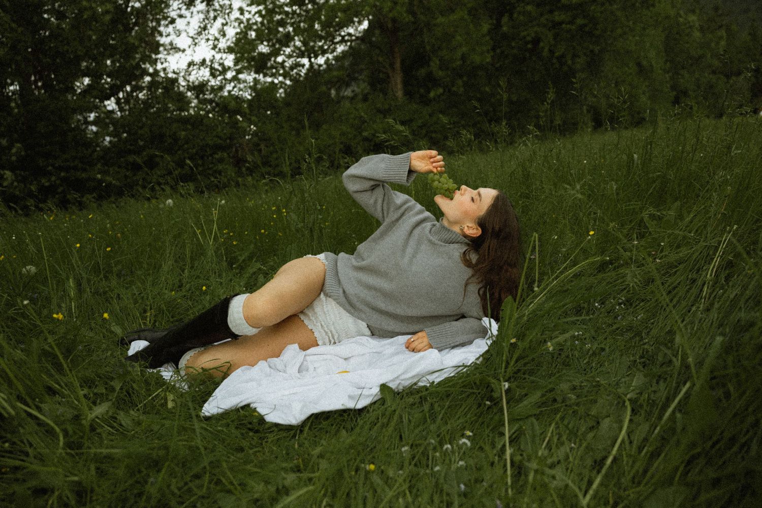Woman in grey hoodie relaxing on white blanket in green grass field outdoors.