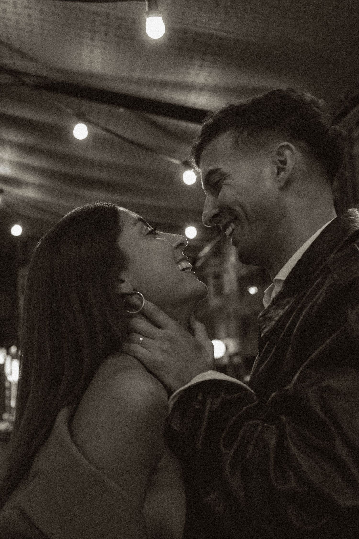A happy couple gazes at each other under string lights in a romantic black and white photo.