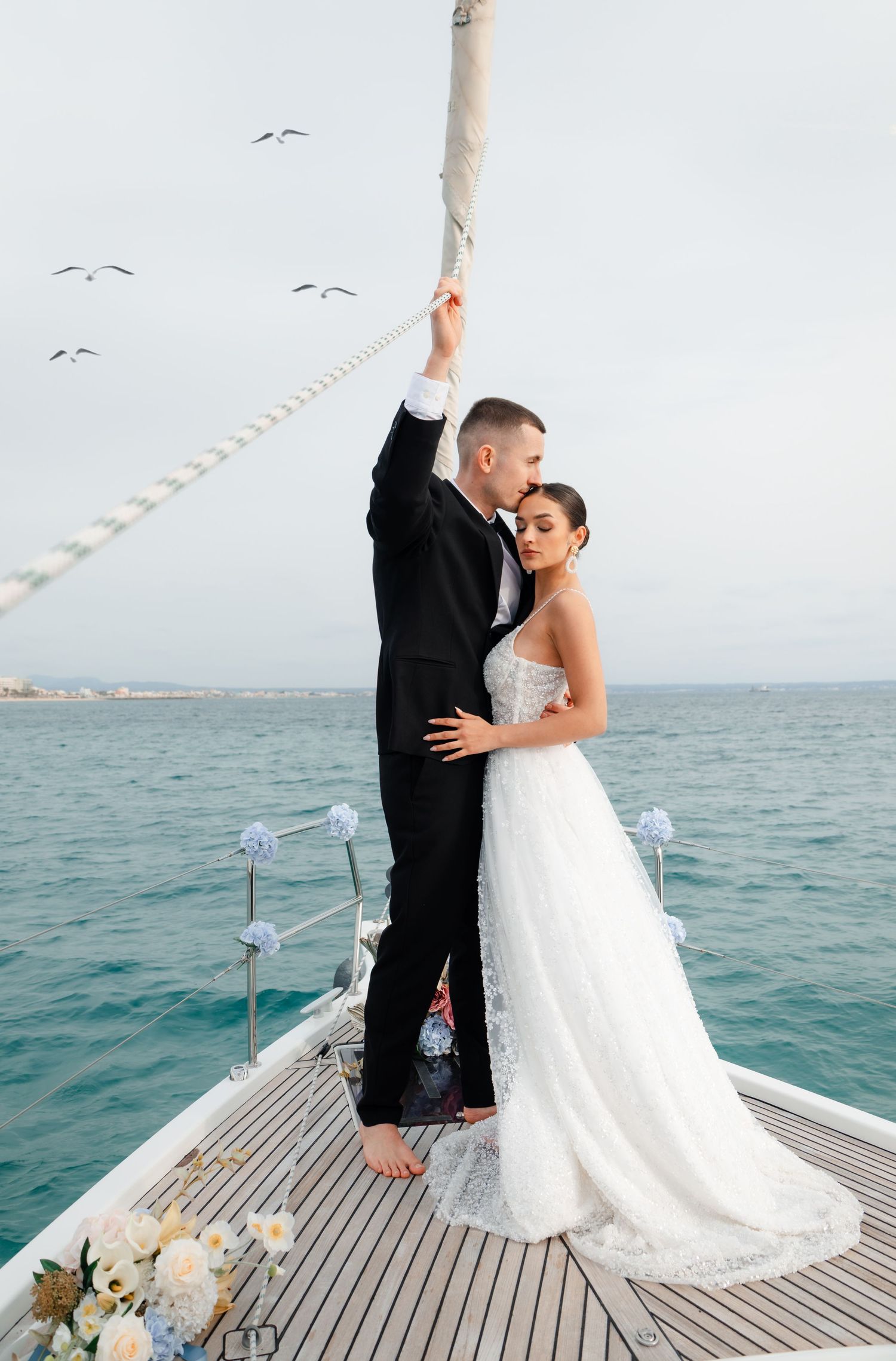 Bride and groom embracing on a sailboat bow with seagulls flying over calm blue ocean waters.