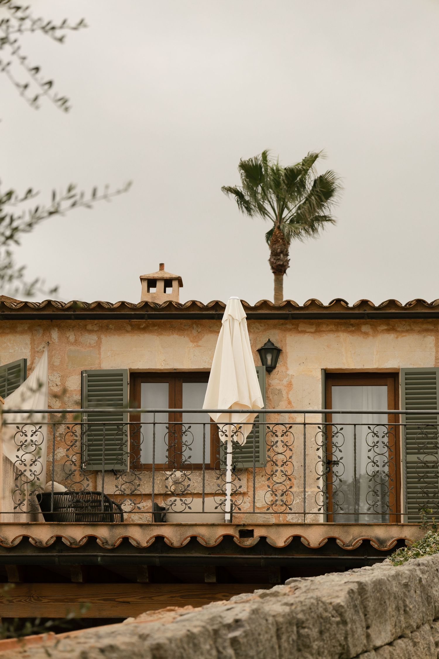 Rustic Mediterranean villa with green shutters, wrought iron balcony, and a palm tree against cloudy sky.