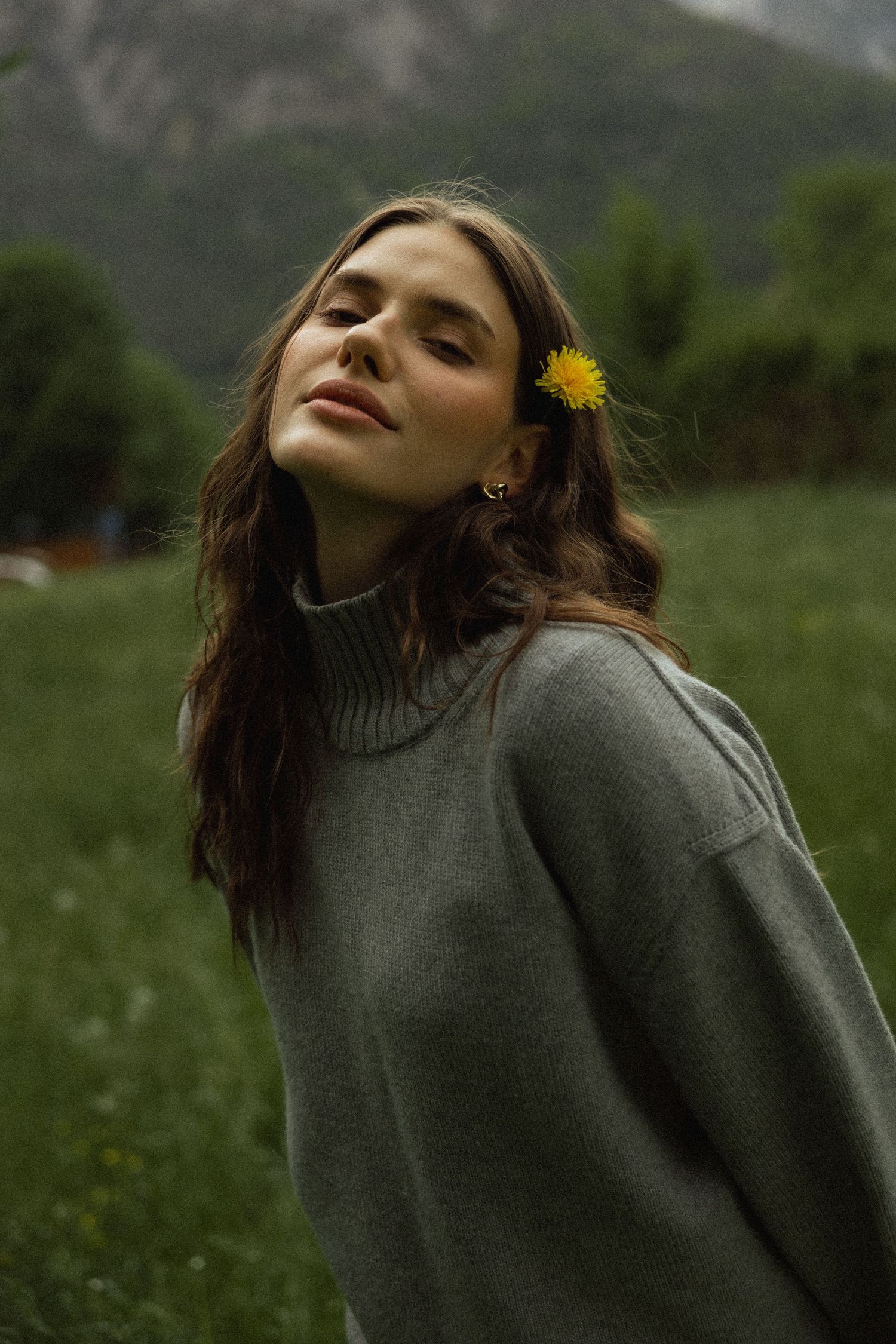 Serene woman in grey turtleneck sweater with yellow flower in hair in mountain field.