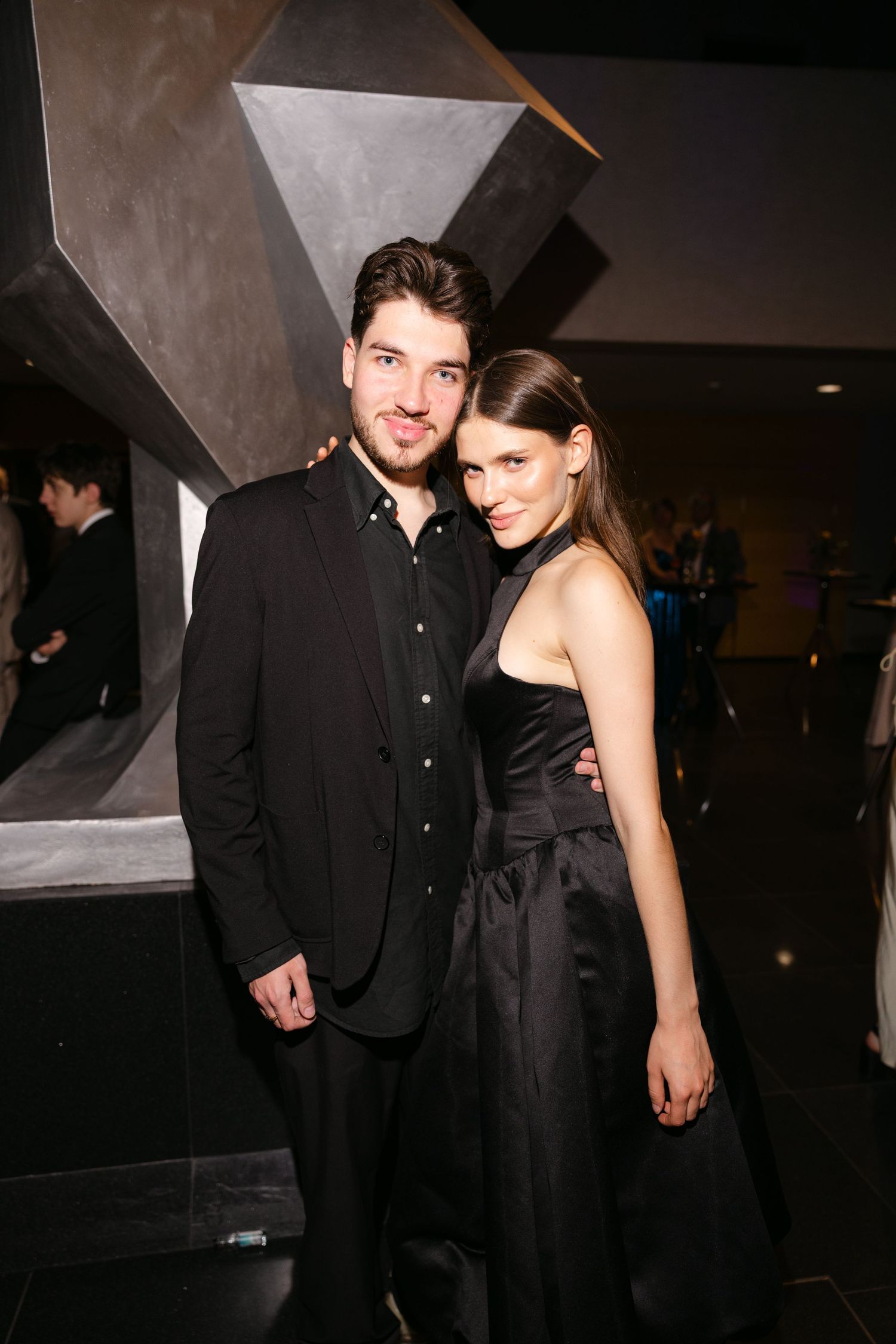 Stylish couple in black formal attire pose together at an upscale evening event.