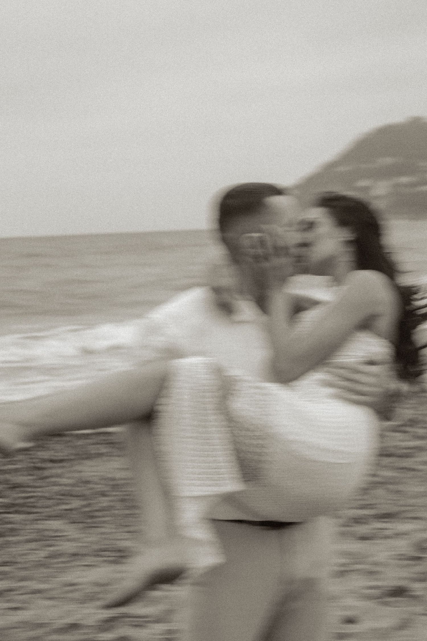 Black and white photo of a couple kissing on a beach, man carrying woman in his arms by the ocean.