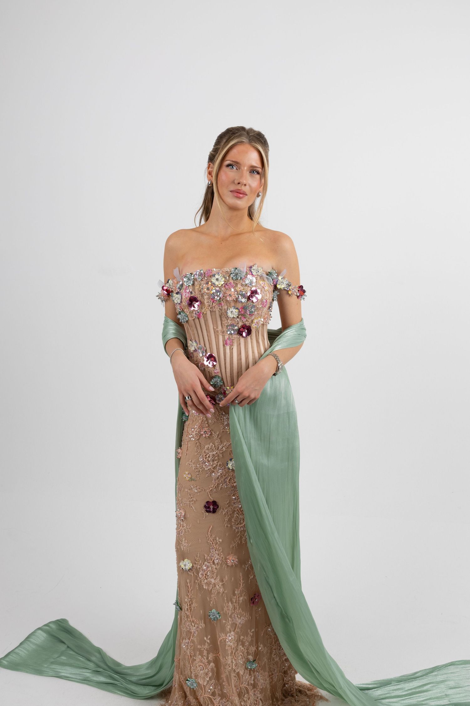 Woman in elegant off-shoulder floral embroidered gown with sage green chiffon overlay on white background.