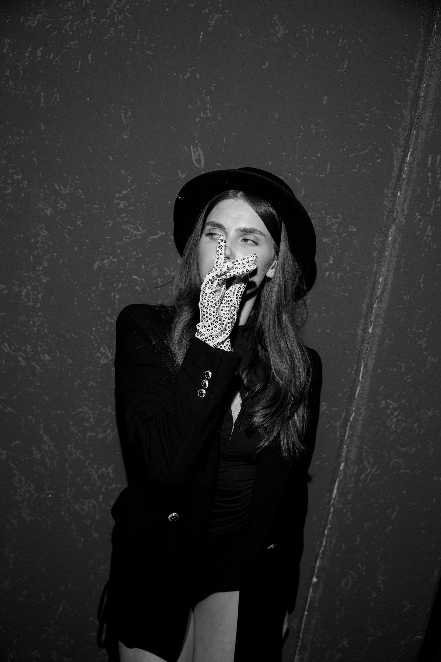 Black and white portrait of a woman in a black hat and blazer covering her mouth with a gloved hand.