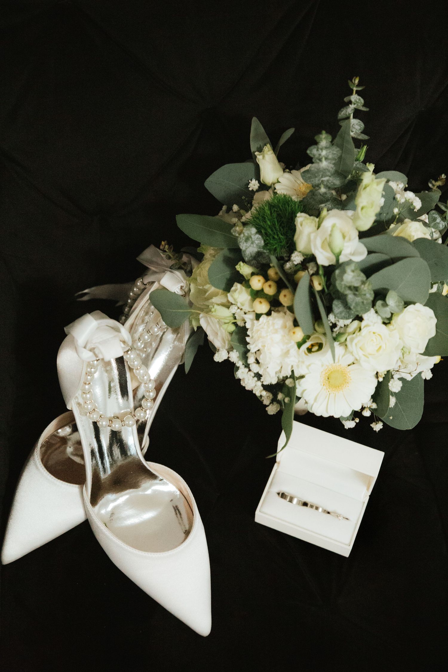 White bridal heels with pearl details beside a white floral bouquet and ring box on dark background.