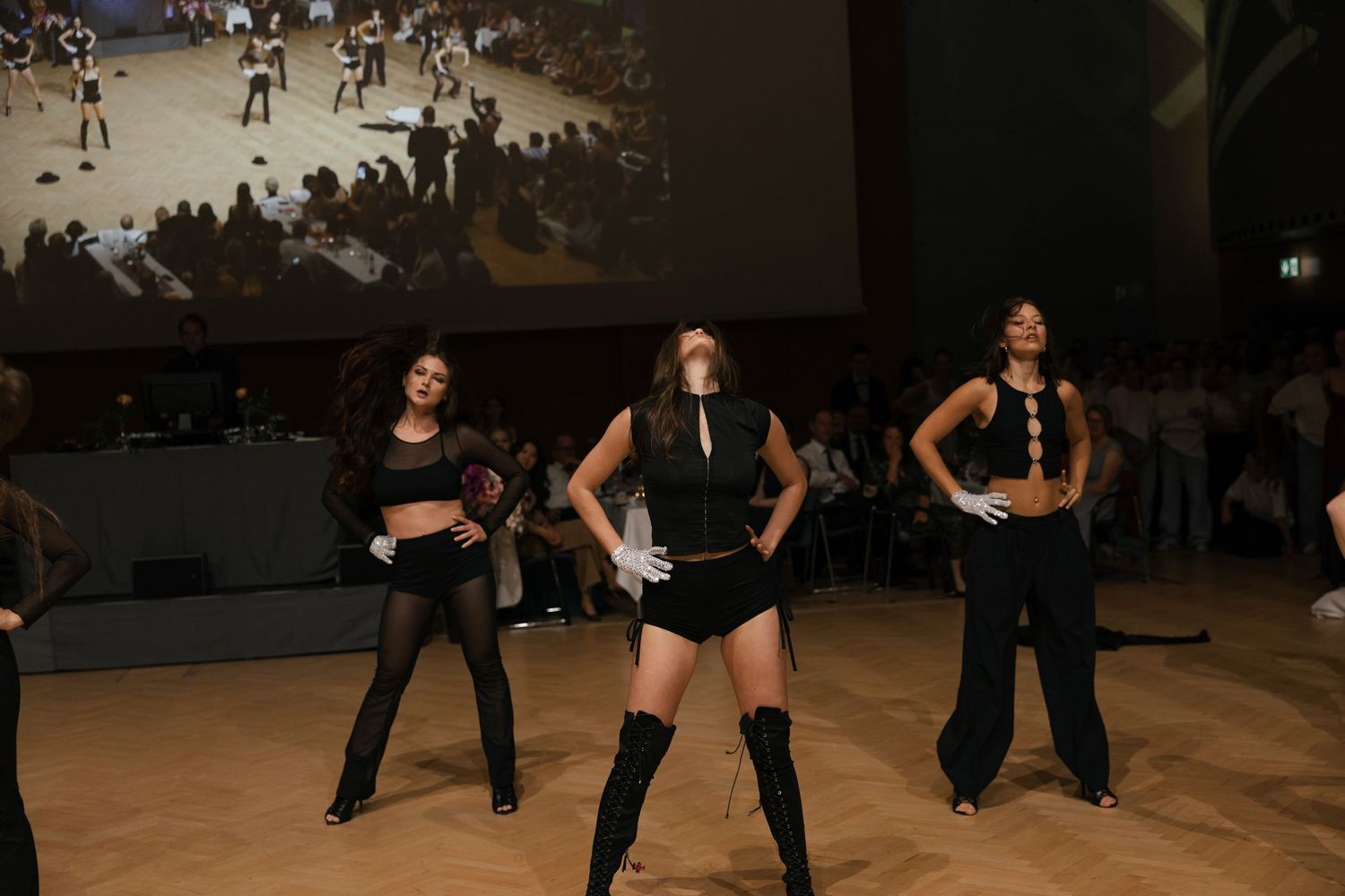 Three dancers in black outfits performing choreography on a stage with large screen backdrop.